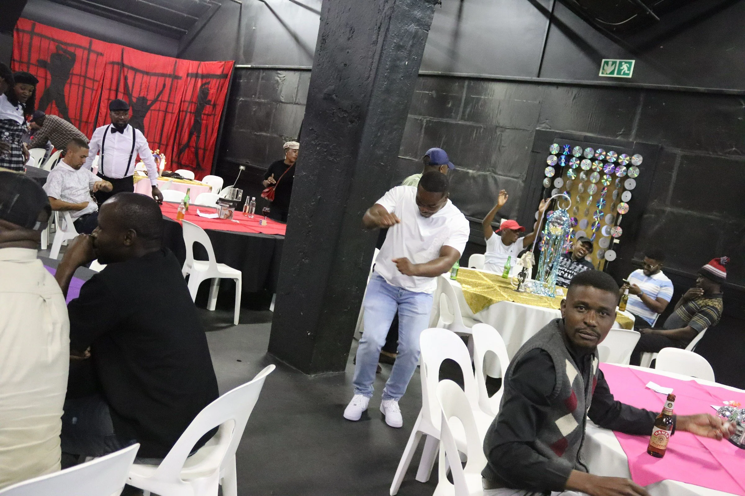 A group of people at a party, some are seated at tables with pink and purple tablecloths, others are standing and dancing, with decorations including a large red fabric backdrop with black silhouettes and a table with colorful CD mirror decor.