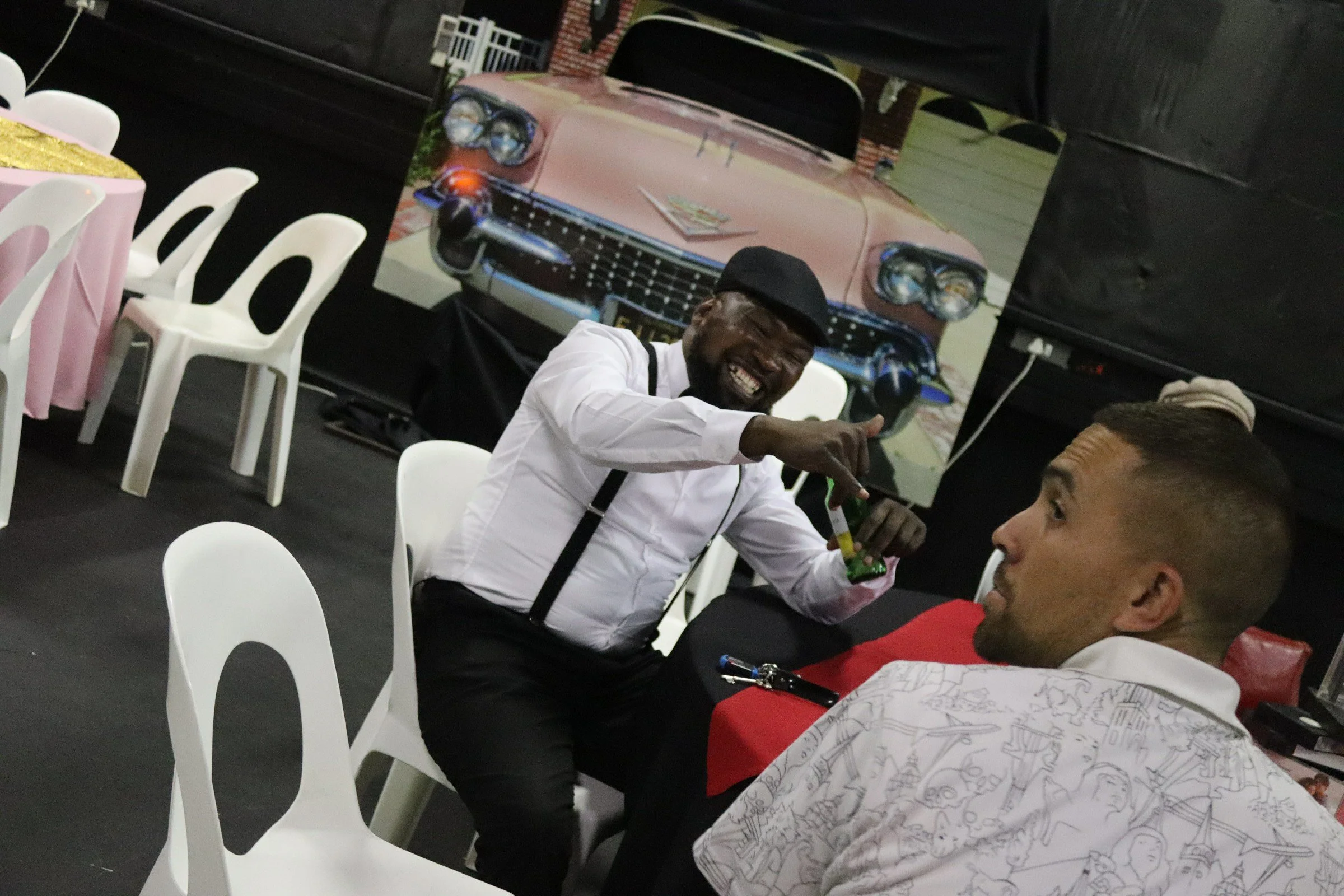 A man wearing a white shirt, black suspenders, and a black hat is sitting at a table, smiling, and pointing with his right hand. On the table, there are some items, including a green snack. In the background, there is a large poster of a vintage pink
