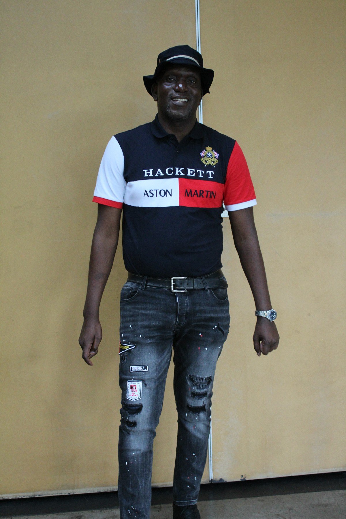 A man standing in front of a beige wall, wearing a black, white, and red Hackett polo shirt, a black hat, distressed jeans, a watch, and a bracelet, smiling at the camera.