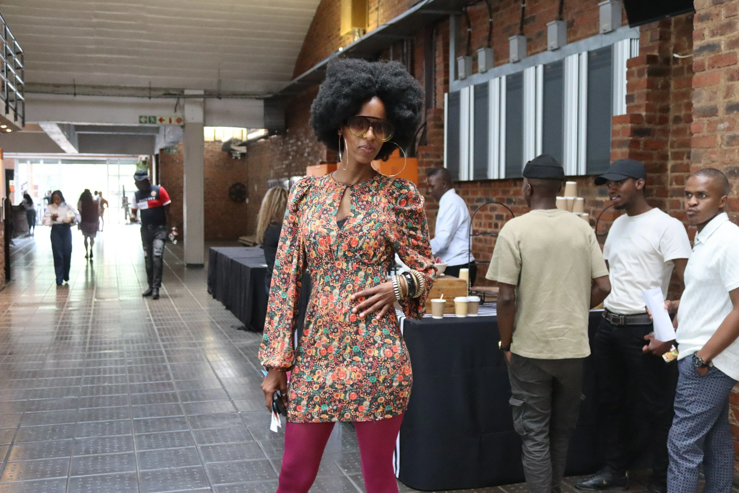 A woman with large afro hairstyle wearing sunglasses, a floral dress, maroon tights, and multiple bracelets on her right wrist, standing indoors with several people in the background.