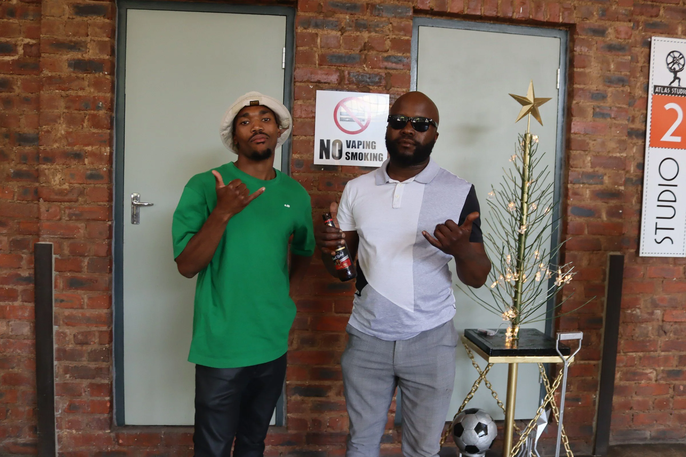 Two men standing in front of a brick wall with a 'No Smoking' sign. One man is wearing a green shirt and a bucket hat, making a hand sign, and the other man is wearing a light-colored shirt with a dark patch on the sleeve, sunglasses, and holding a b