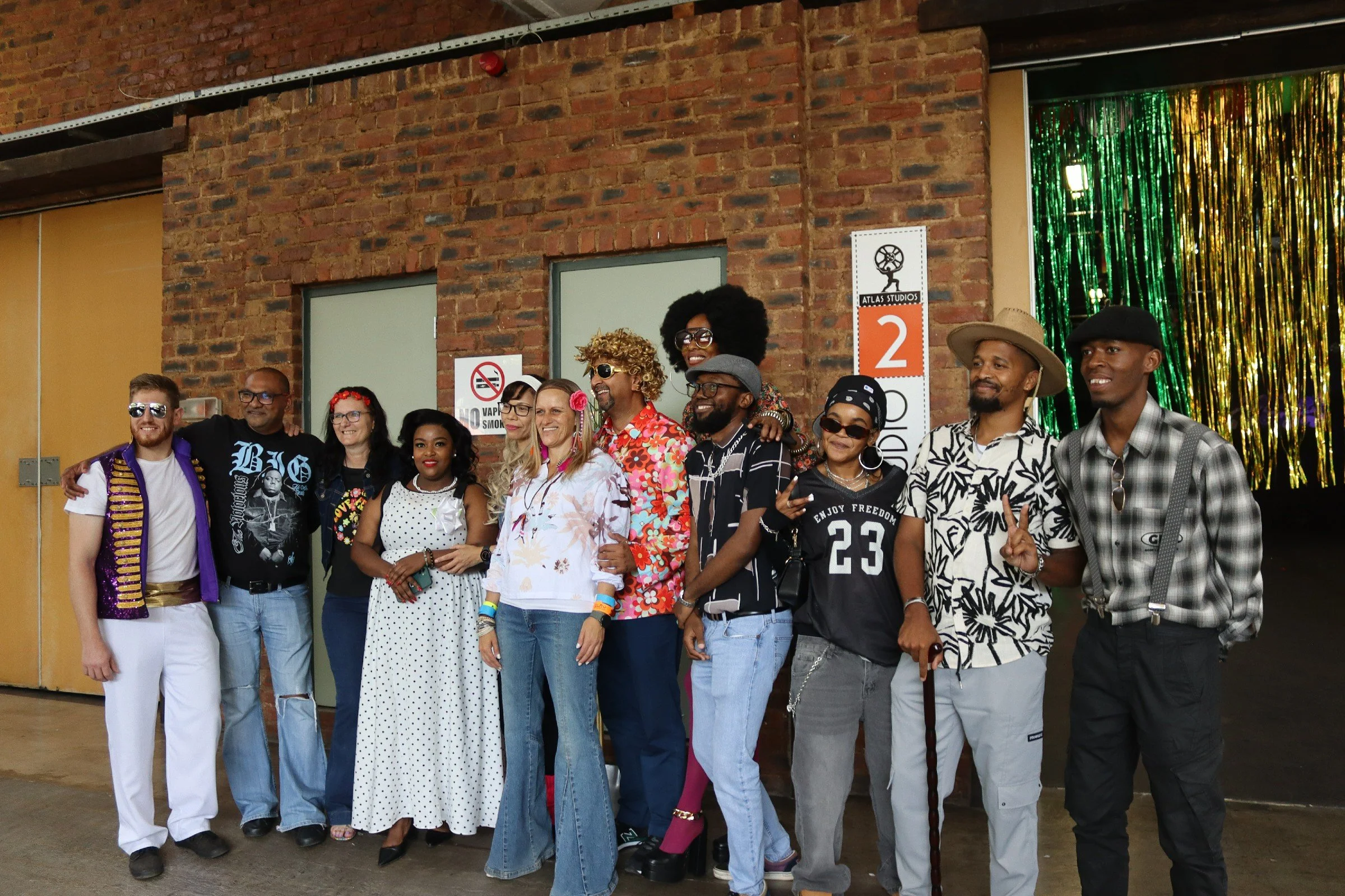 A group of diverse people posing together indoors in front of a brick wall, with colorful decorations and a sign reading 'Atlas Studios' behind them.