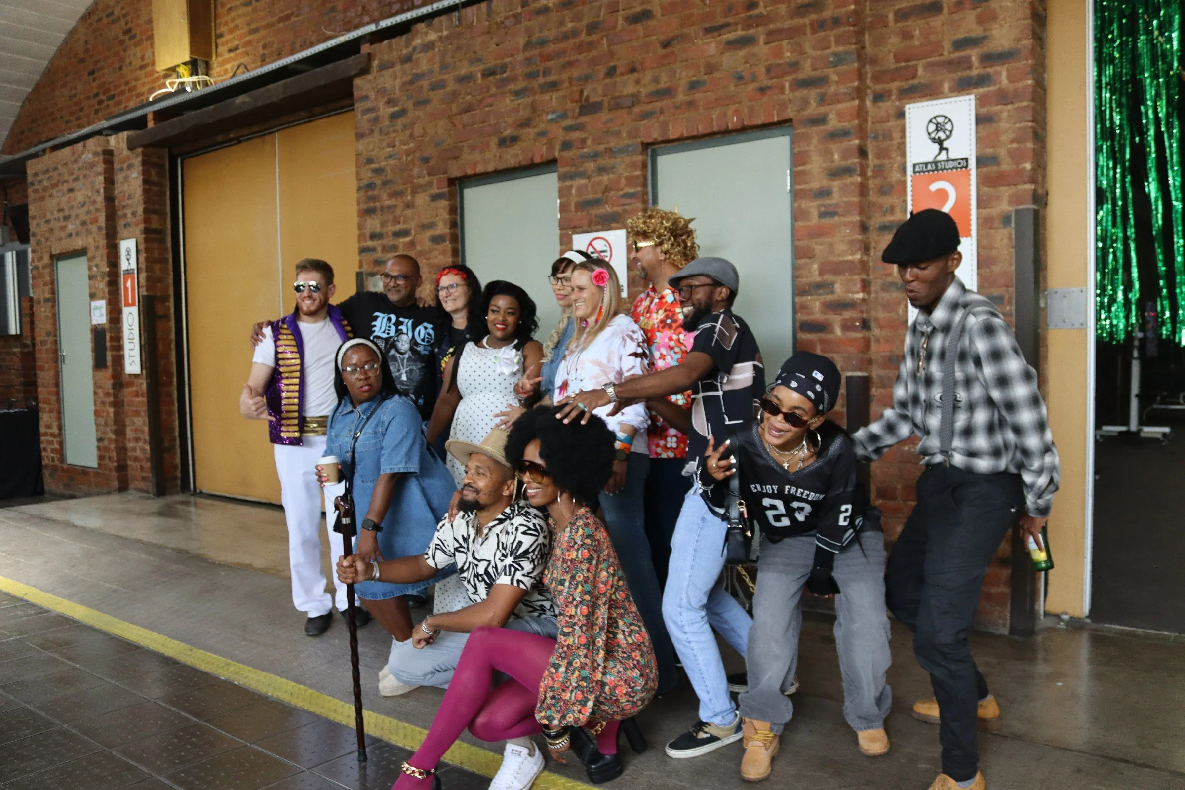 Group of diverse people posing for a photo outside a studio location. They are smiling and dressed in colorful, casual and vintage-style clothing. Some are standing, while others are seated or crouching in front. The background features a brick wall 