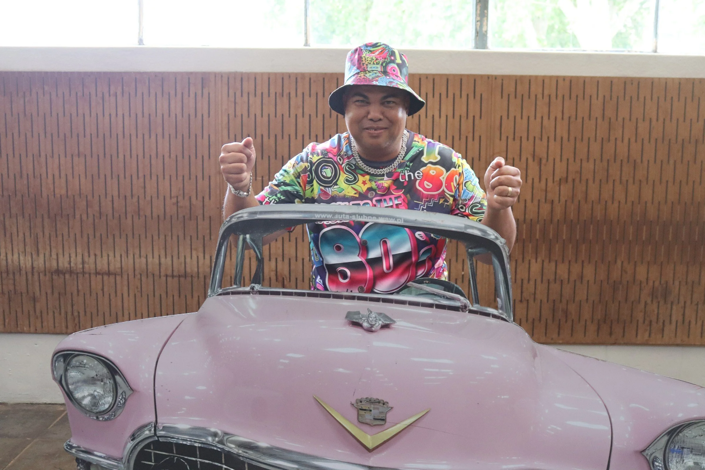 A man wearing a colorful shirt and hat sitting in a small pink toy Cadillac. The car has a emblem on the hood and is in an indoor setting with a wooden paneled wall behind him.