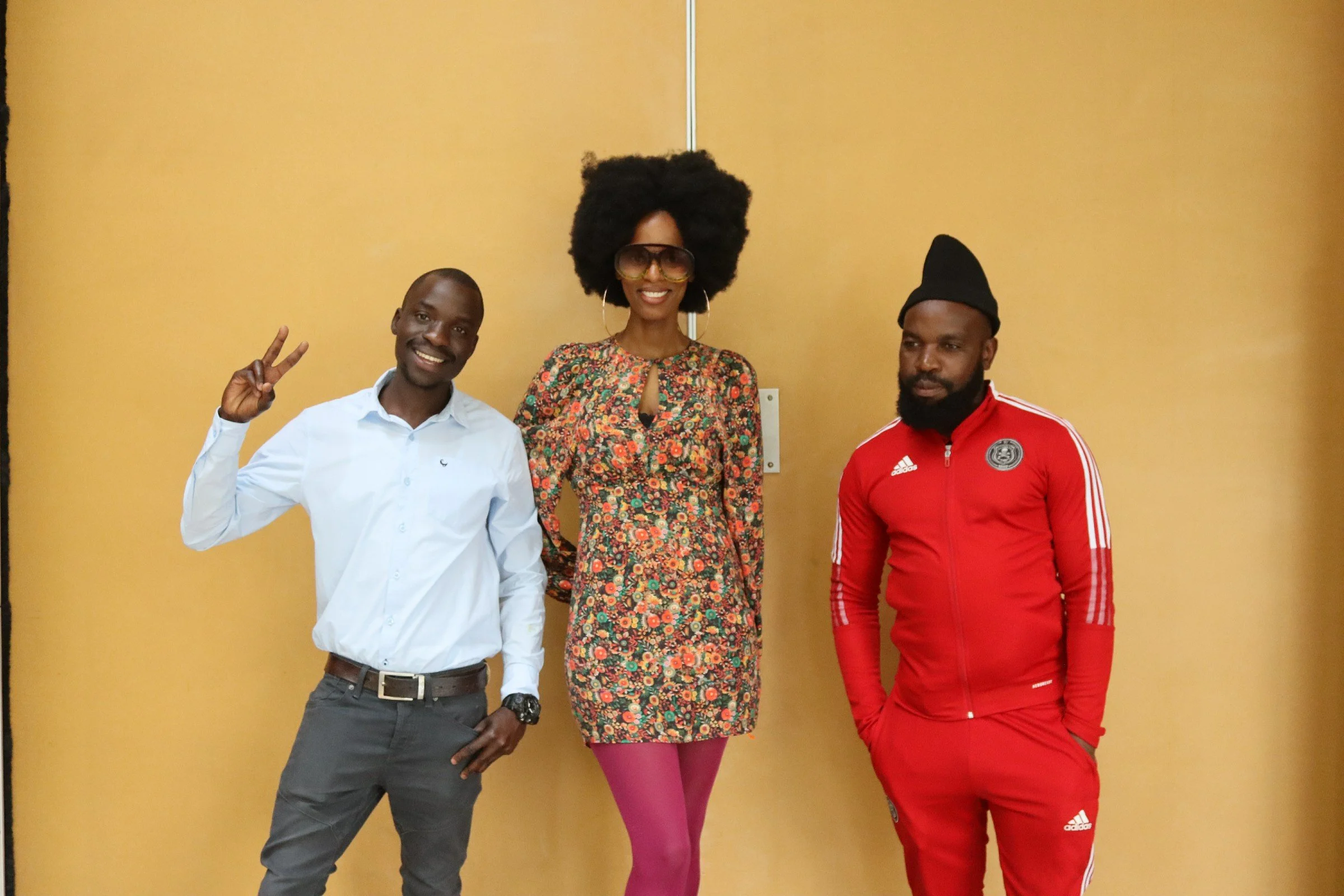 Three people standing in front of a yellow wall. The person on the left is a man wearing a white shirt and gray pants, making a peace sign with his right hand. The person in the middle is a woman with an oversized afro hairstyle, large sunglasses, a 