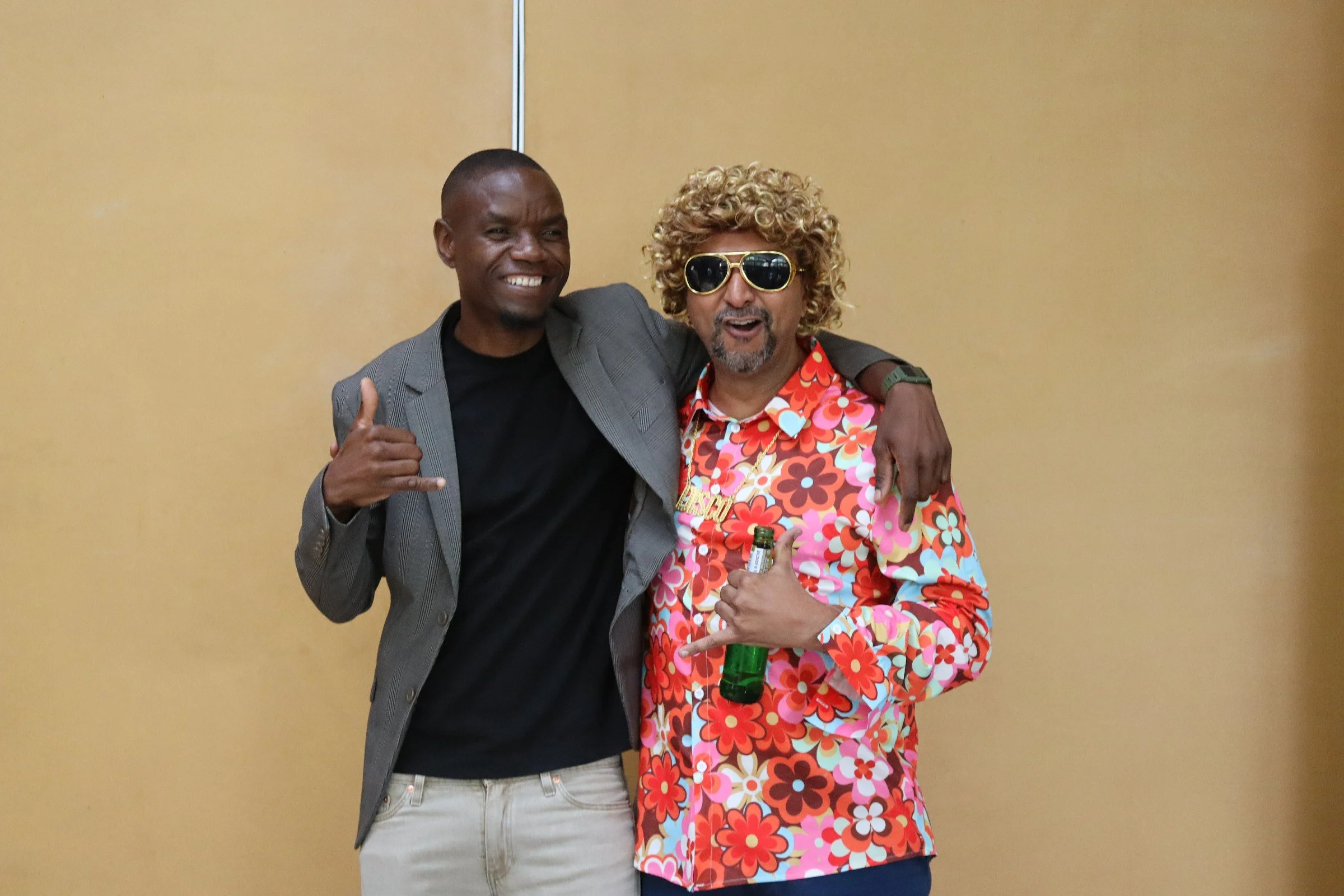Two men standing arm-in-arm smiling at the camera in front of a plain yellow wall. One man is wearing a charcoal blazer and white pants, giving a thumbs-up. The other man is dressed in a colorful, floral-patterned shirt and dark sunglasses, holding a