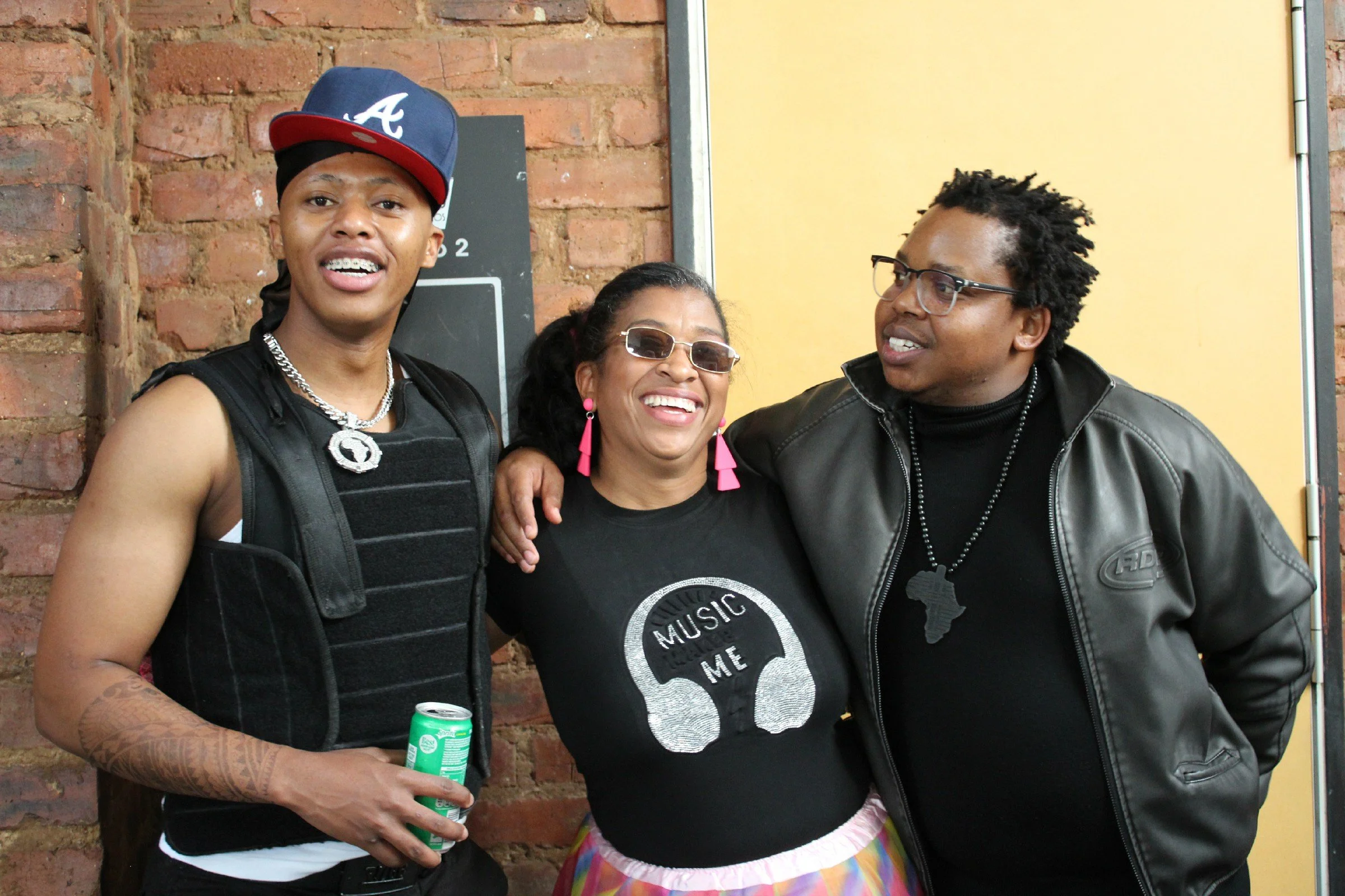 Three people smiling and posing together in front of a brick wall and a yellow door. The person on the left is wearing a black cap with red and white accents, braces, a silver chain, a black vest, and has tattoos on their arm. The middle person is we