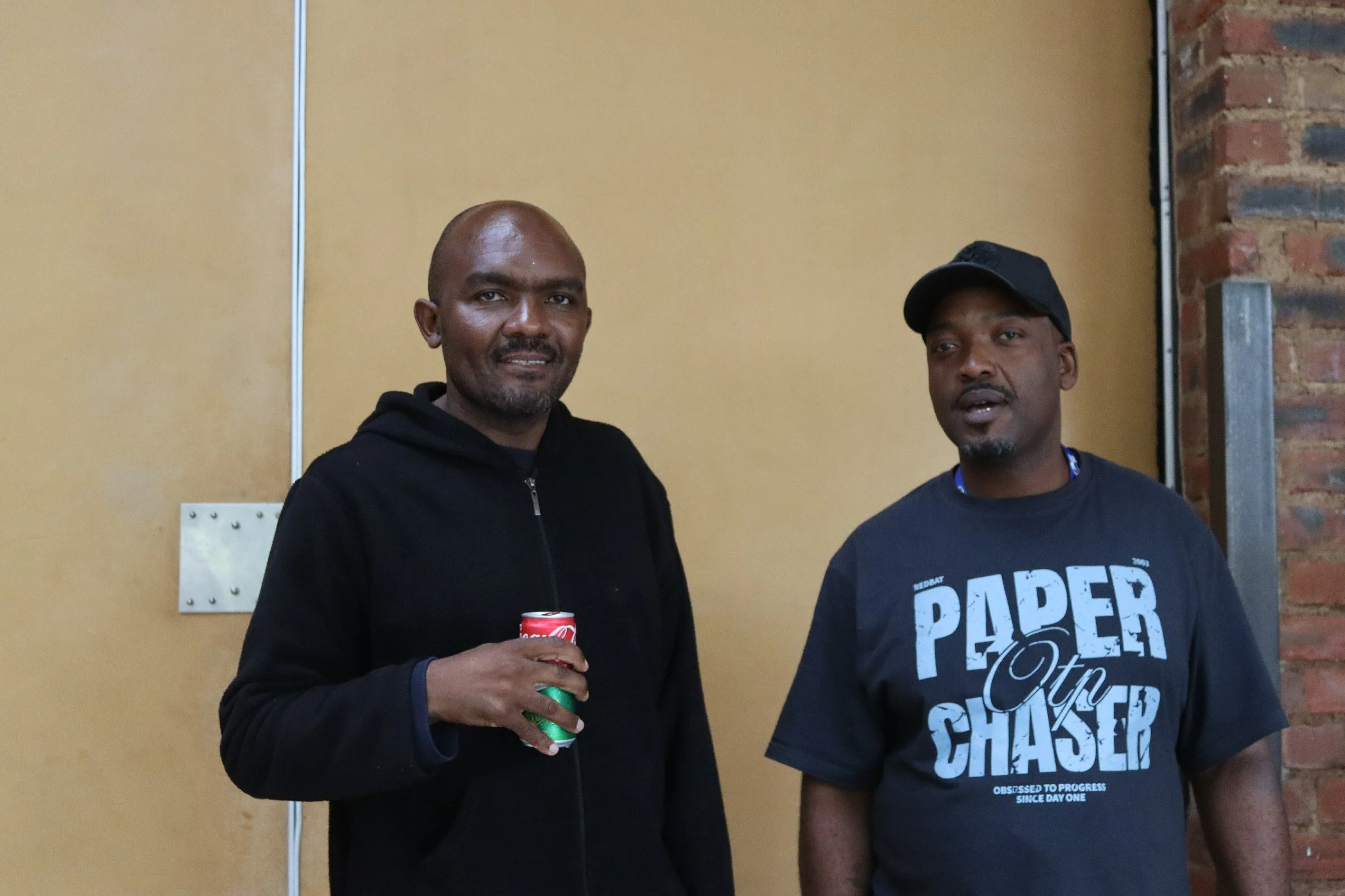 Two men standing indoors in front of a beige wall and a brick column. The man on the left is bald with a beard, wearing a black hoodie, and holding a can of soda. The man on the right is wearing a black cap, a dark T-shirt with text, and has an open 
