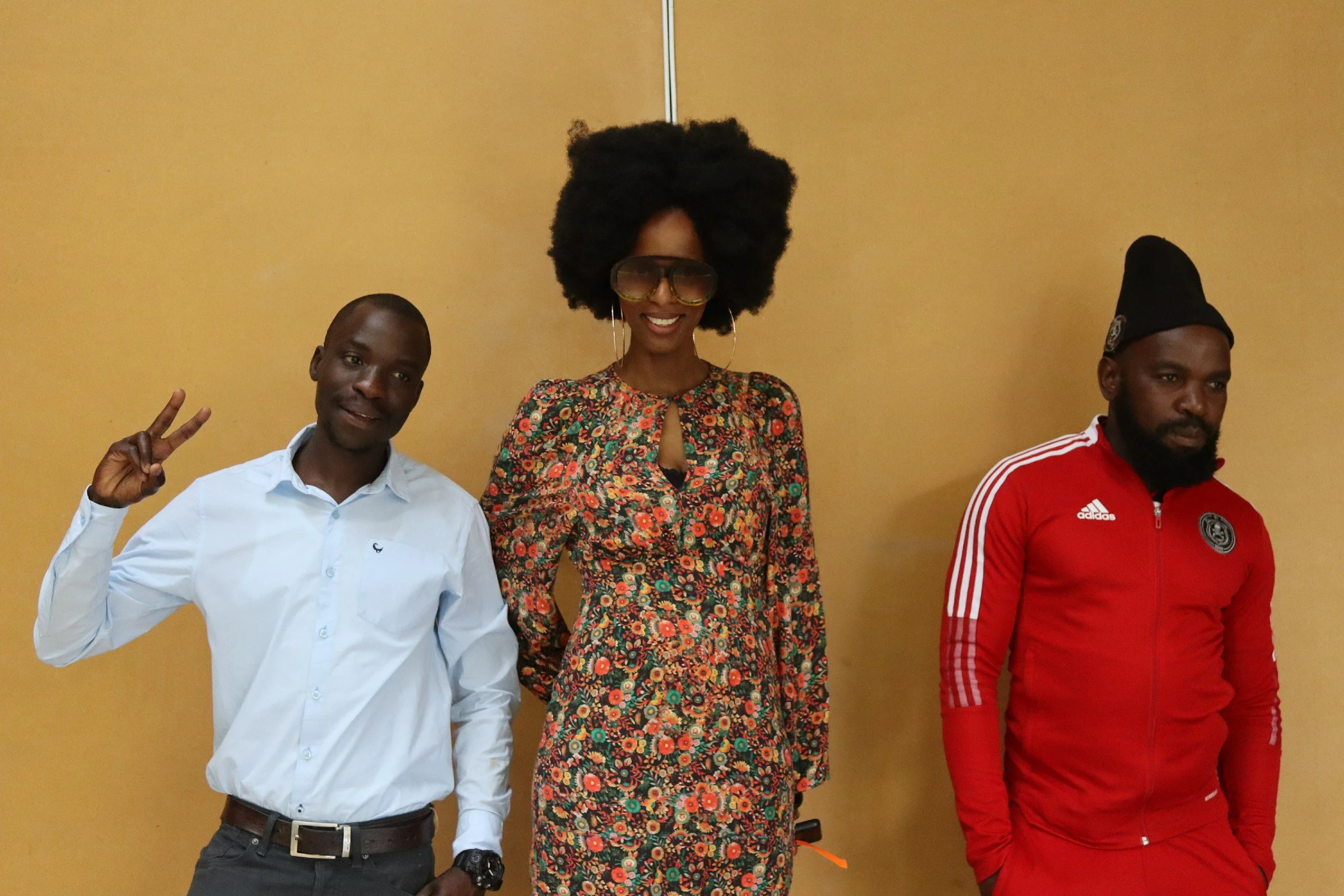 Three individuals standing against a yellowish wall. The person on the left is making a peace sign with their hand, wearing a white shirt, and black pants. The person in the middle is wearing large glasses, hoop earrings, a floral dress, and has big 