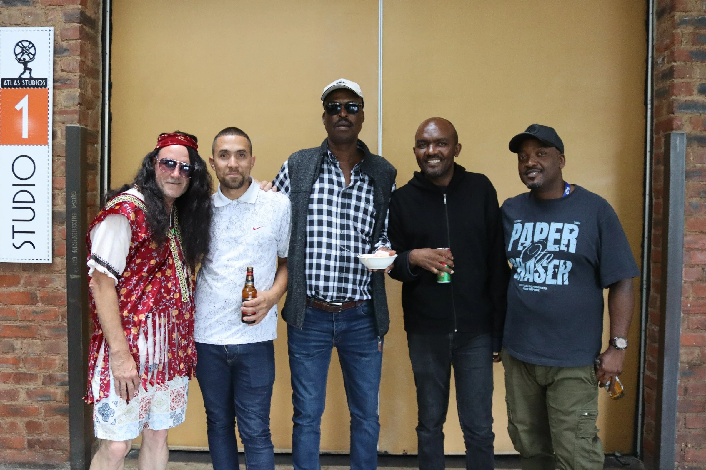 Five men and one woman standing together in front of a beige wall with brick accents, posing for a group photo. One person is holding a bowl and a drink, and another is holding a beer bottle.