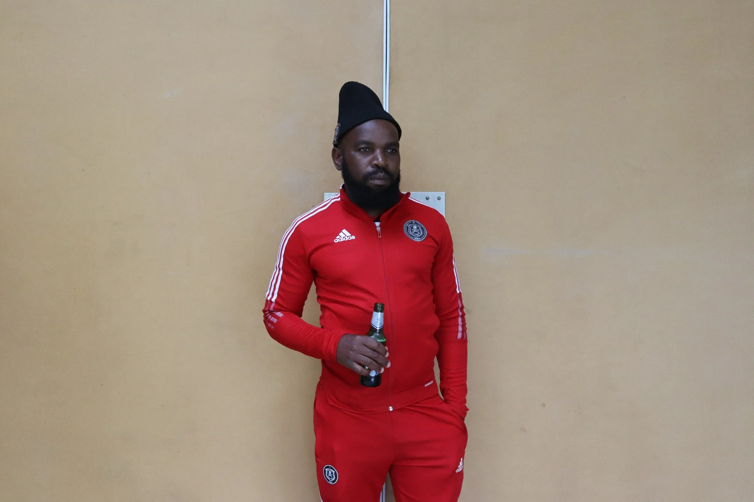 A man with dark skin, a beard, and a serious expression, wearing a black hat and a red Adidas sports jacket with a team logo, holding a beer bottle, standing against a beige wall.