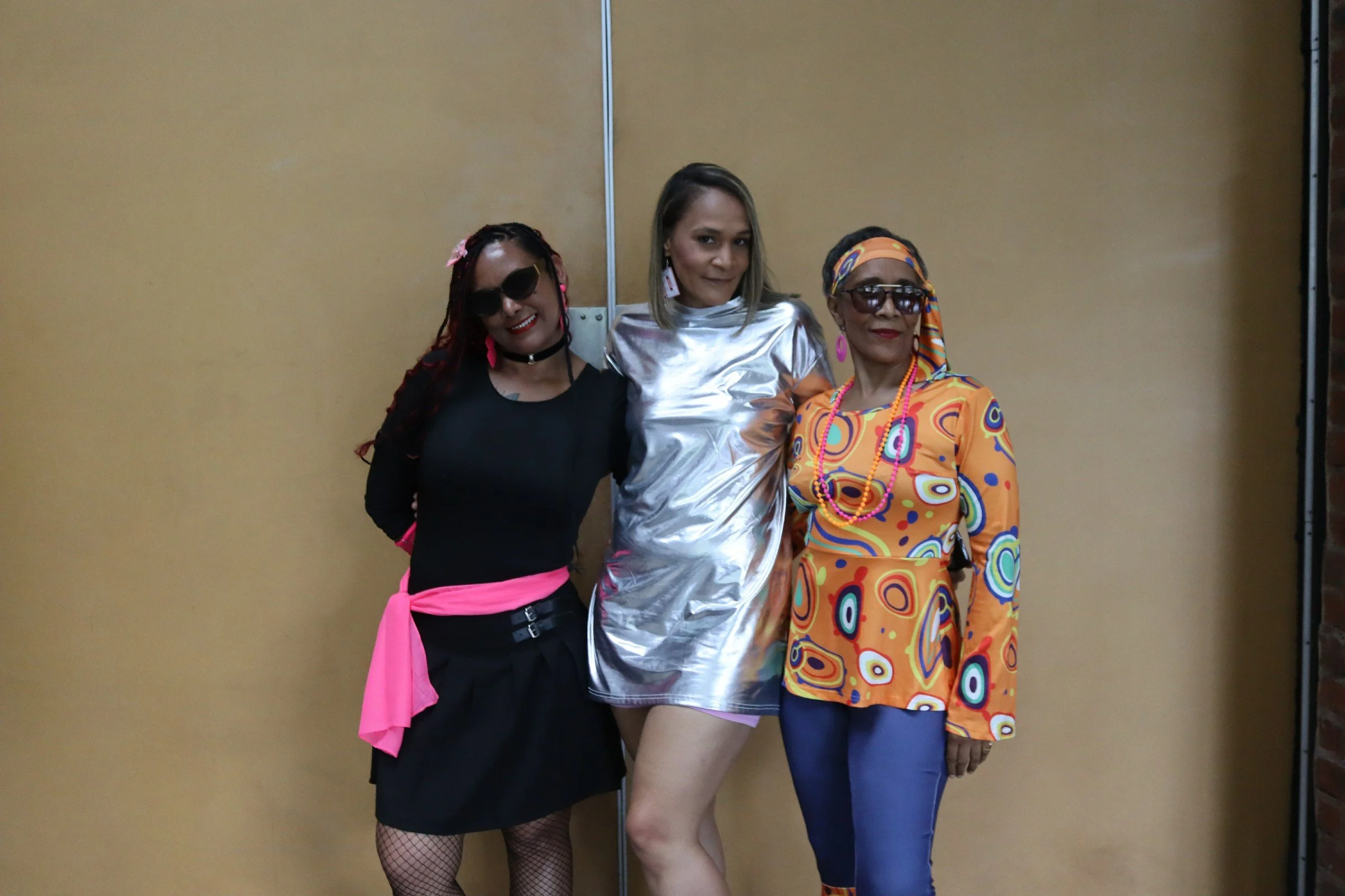 Three women dressed in colorful and vibrant outfits, standing close together, smiling, in front of a plain wall.
