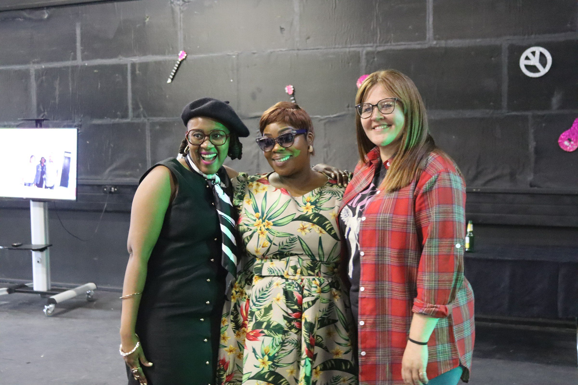 Three women standing close together indoors, smiling, with a dark wall and decorative wall hangings behind them.