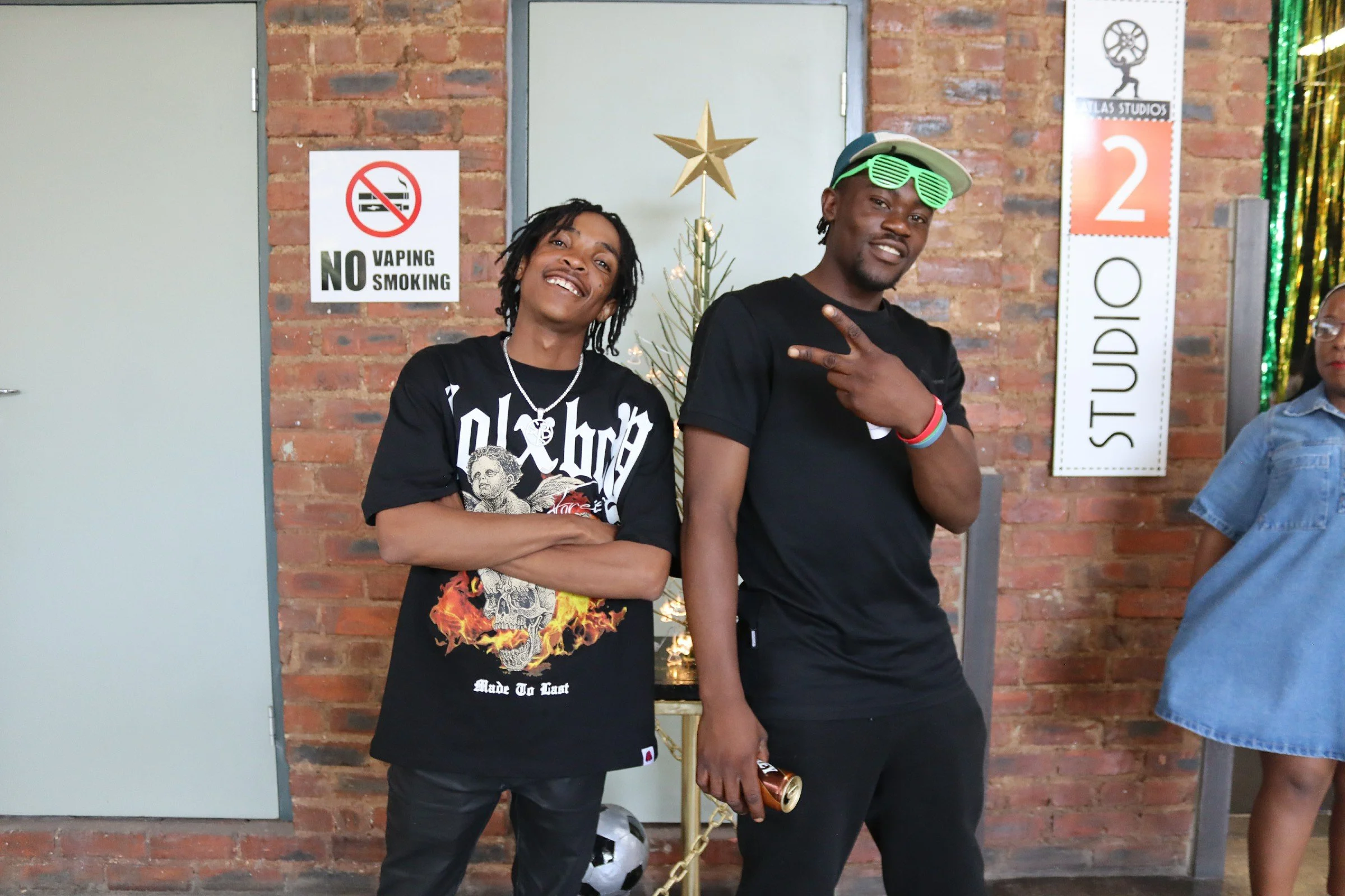 Two men standing in front of a brick wall, smiling. One has dreadlocks and is wearing a black graphic T-shirt. The other is wearing a baseball cap and green shutter shades. There is a no vaping or smoking sign and a decorative gold star on the wall b
