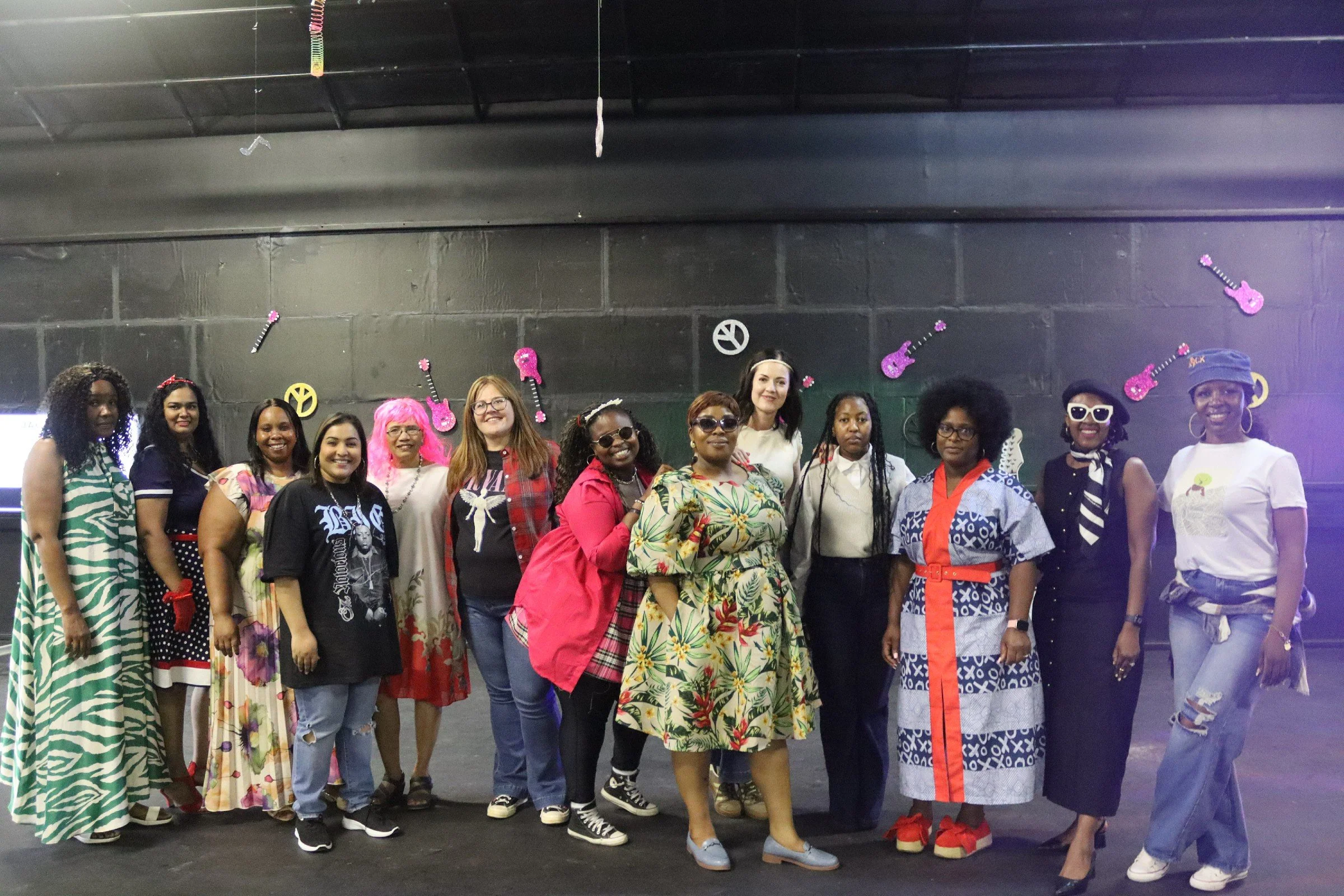 A group of women posing together in a room decorated with pink guitar and peace sign cutouts on a black wall. The women are wearing colorful and patterned outfits, some with accessories like glasses and headbands, smiling for the photo.