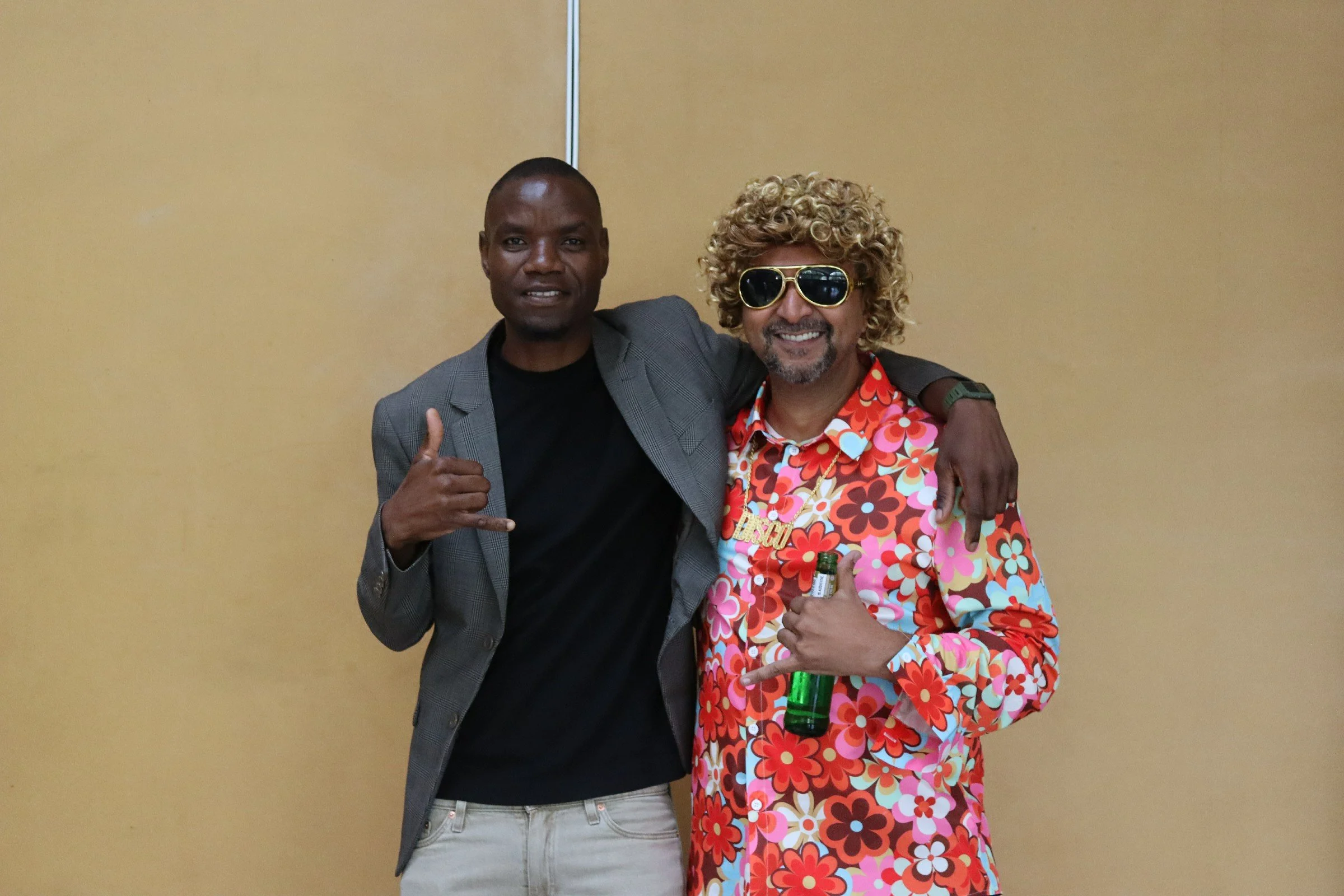 Two men posing together indoors, one with a gray plaid blazer, black shirt, and white pants making a thumbs-up, the other with curly hair, sunglasses, a colorful floral shirt, and holding a green bottle.