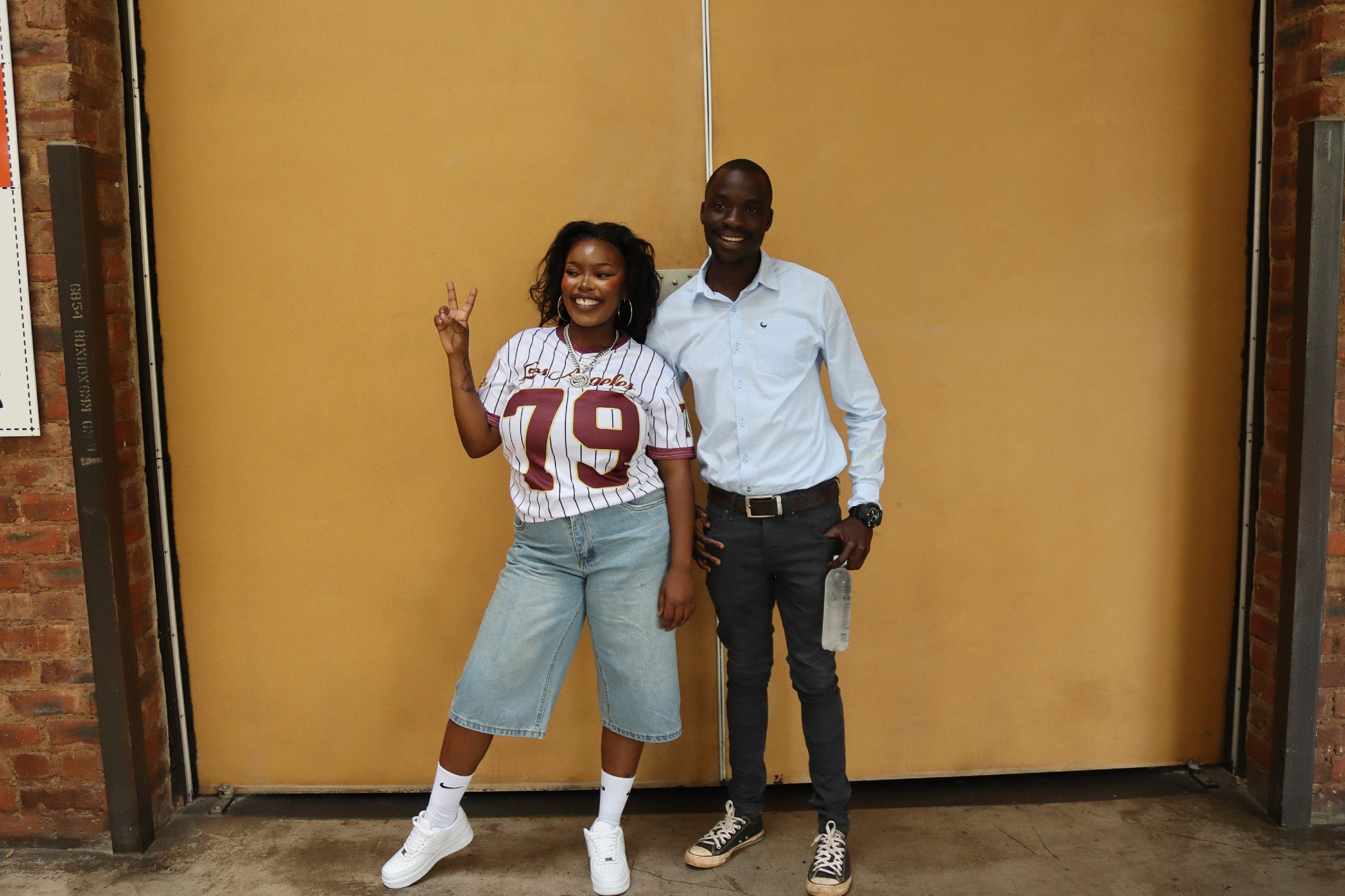Two smiling people standing in front of a yellow partition wall. The woman on the left is wearing a white sports jersey with red and yellow lettering, light blue jeans, white socks, and white sneakers. She has curly dark hair, hoop earrings, and is m