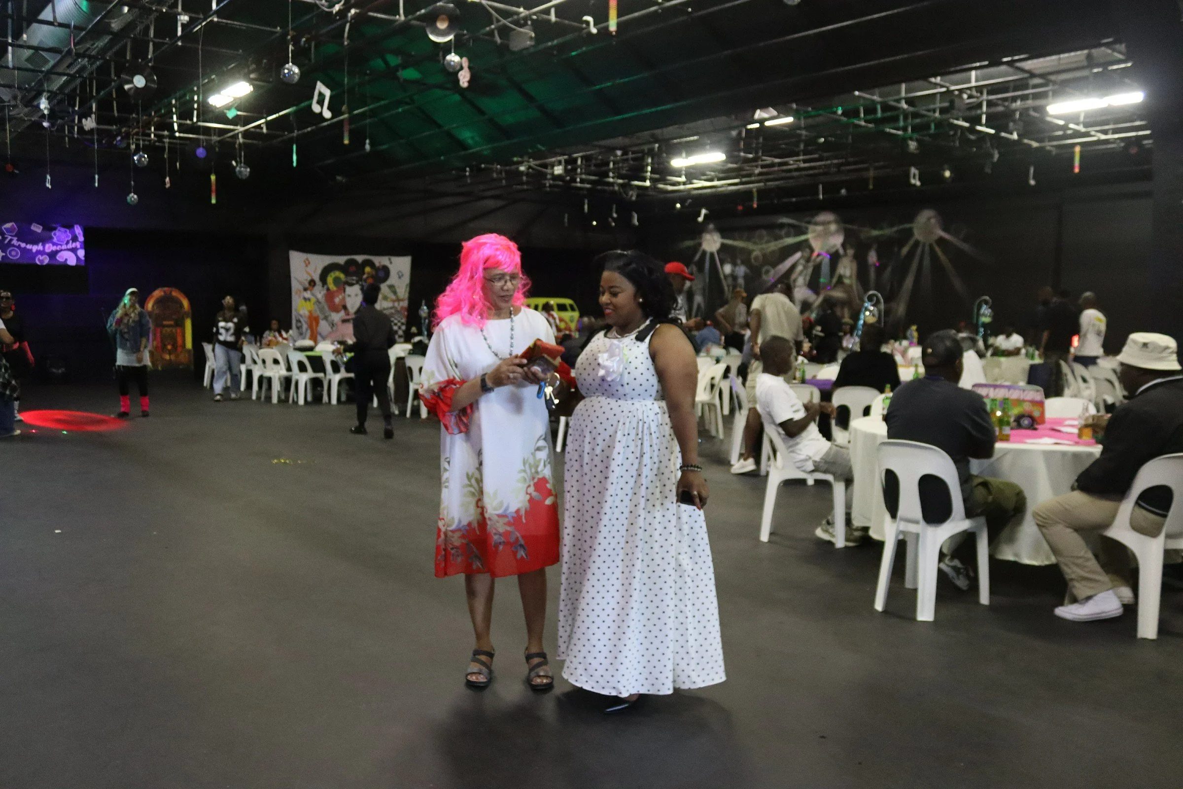 Two women, one with pink hair and glasses wearing a white dress with floral and red accents, and the other woman with black hair in a white dress with black polka dots, are standing and talking at a social gathering or party. The background shows mul