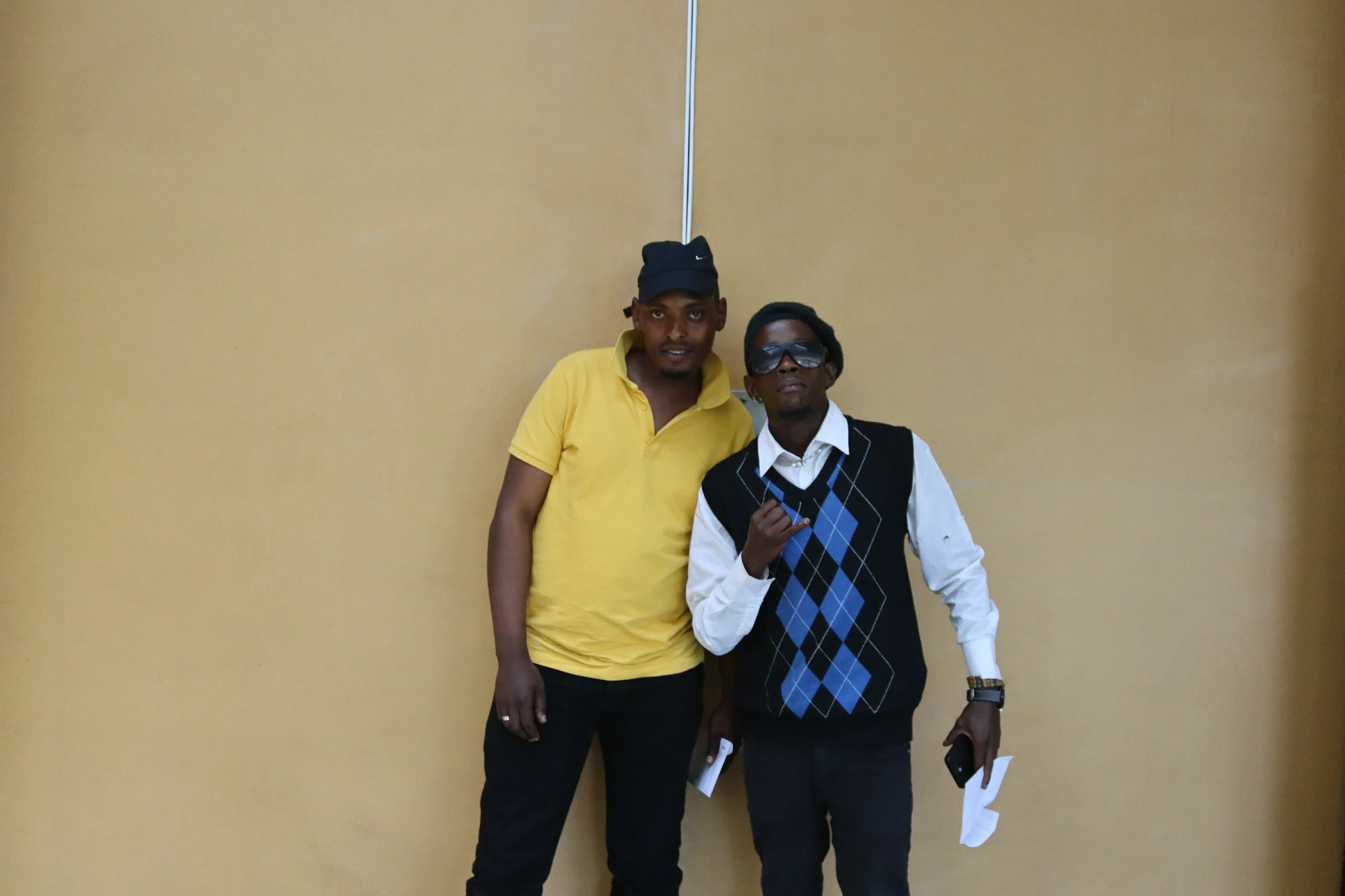 Two men standing against a yellow wall, one wearing a yellow polo shirt and cap, the other in a white shirt with a black and blue argyle vest, sunglasses, and holding papers and a phone.
