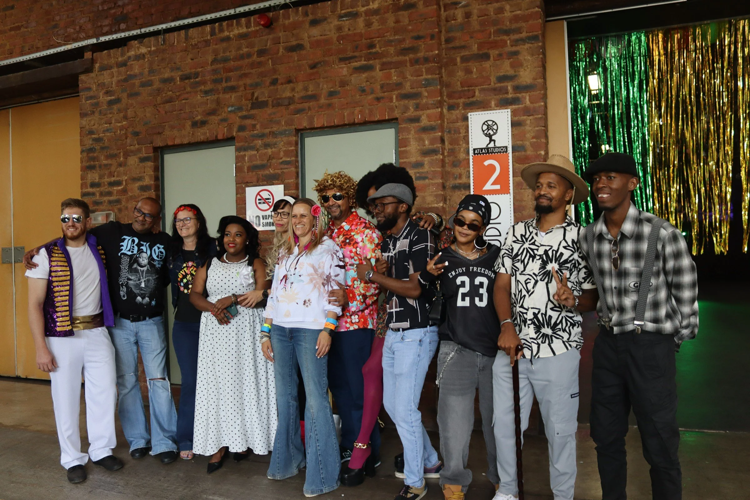 A group of twelve diverse people standing in front of a brick wall and colorful metallic curtains, posing for a photo and smiling.