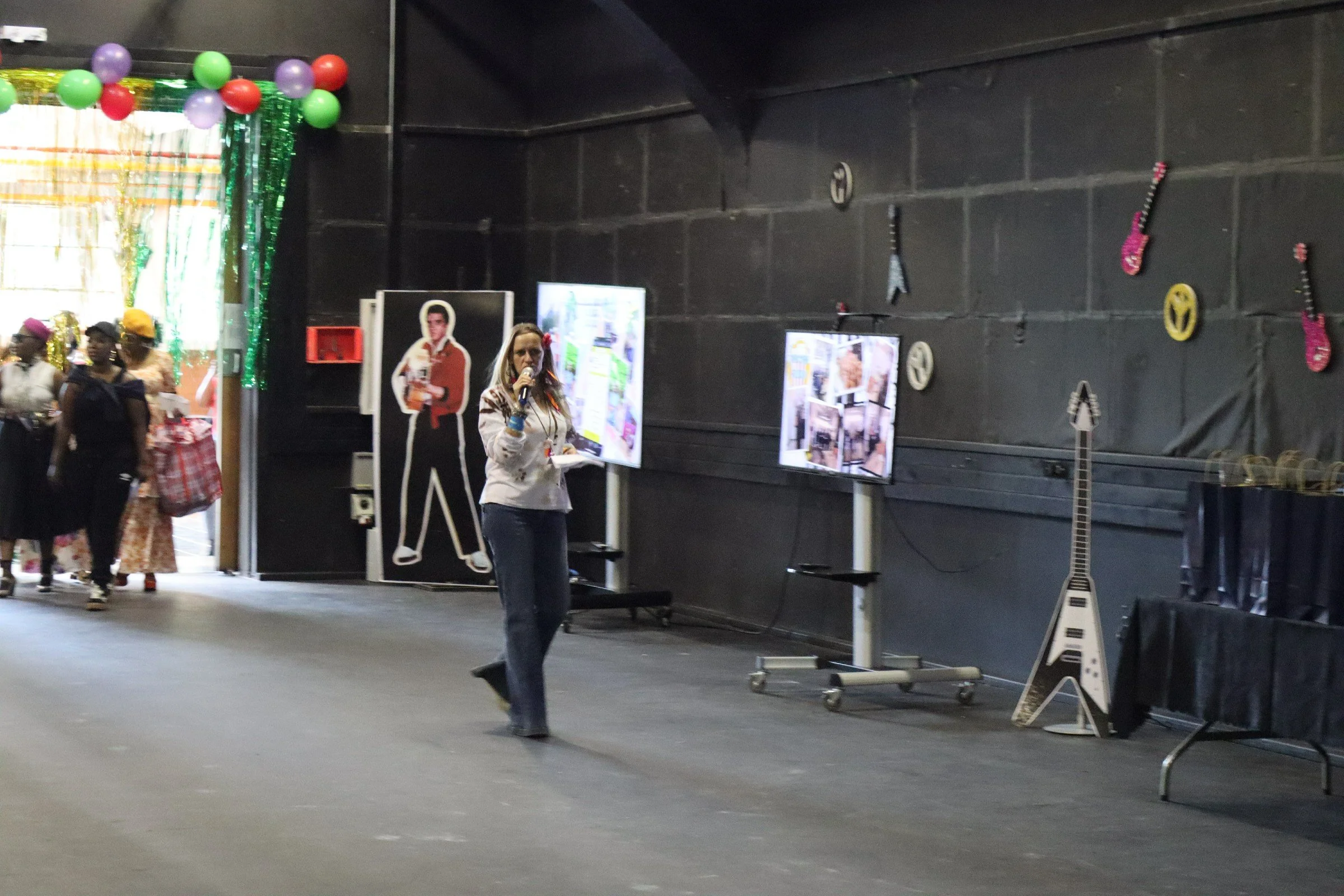 Indoors at a decorated celebration venue with balloons, a woman speaking into a microphone, and a cardboard cutout of a person holding a drink. There are display boards and colorful guitars on the wall.