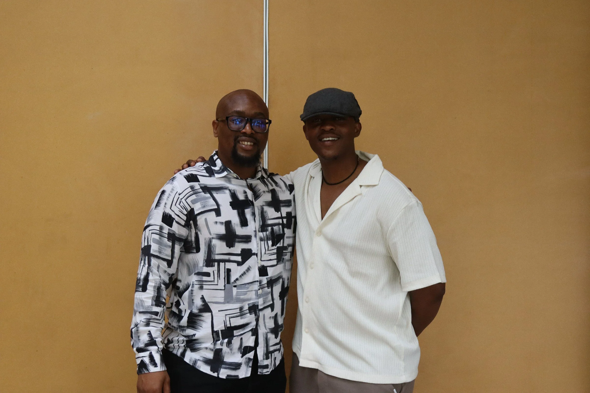 Two men standing together in front of a beige wall, smiling and posing for the photo. The man on the left is wearing glasses and a black and white patterned shirt, and the man on the right is wearing a gray cap and a white shirt.