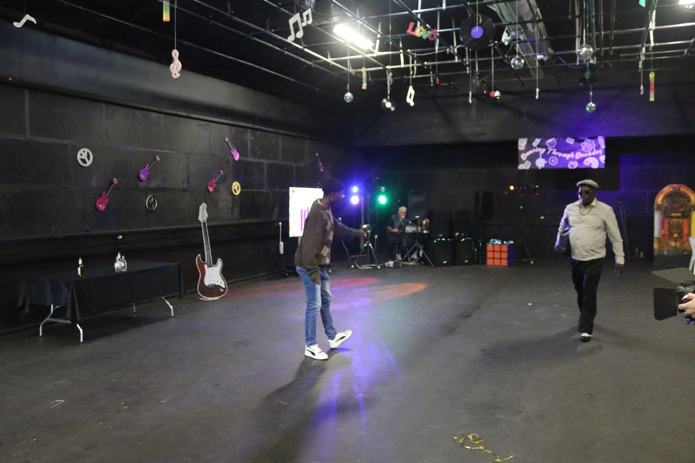 An indoor stage with musical decorations, including guitars and peace signs, with people standing and a DJ setup in the background.