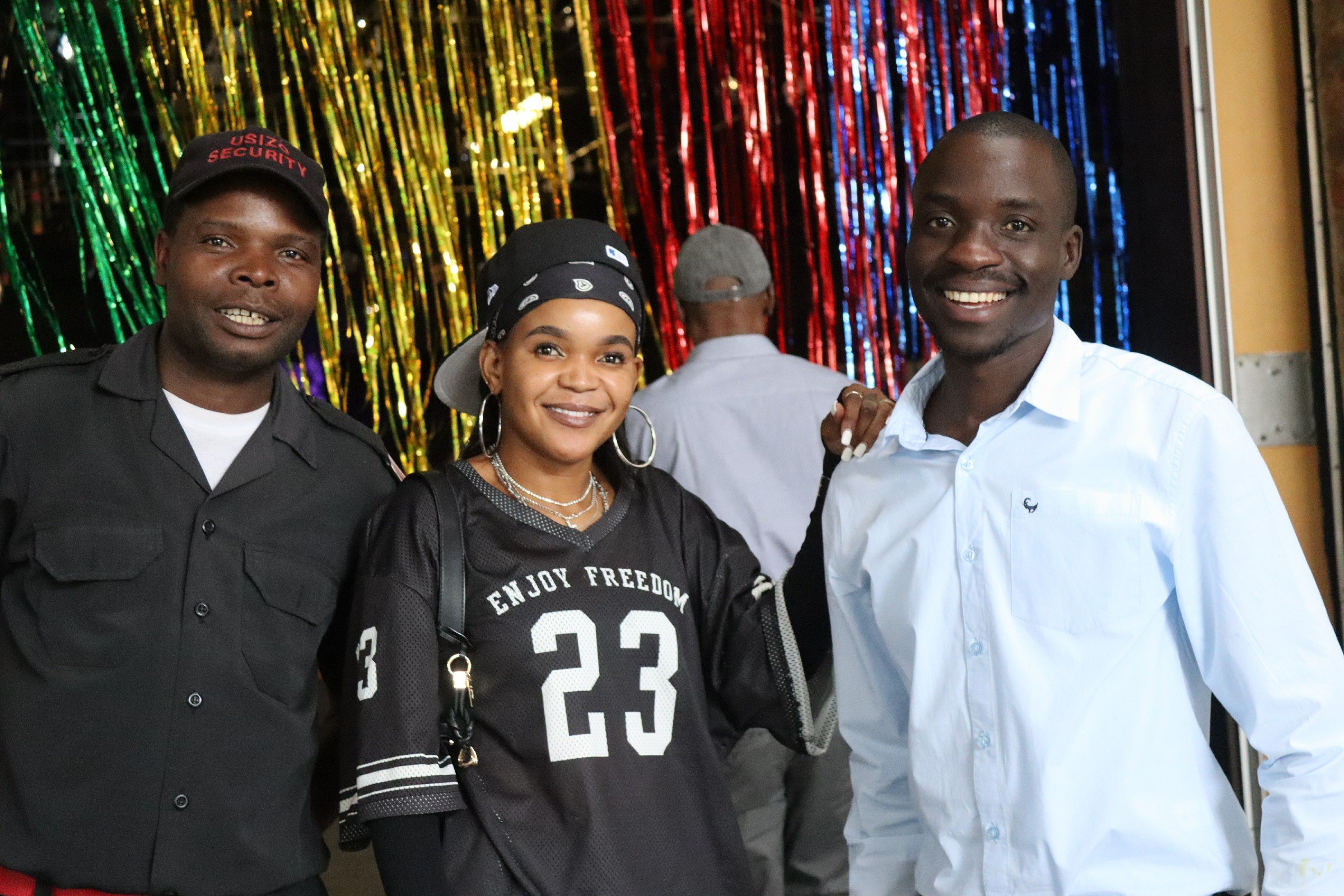 Three smiling people posing together at an event with rainbow-colored metallic streamers in the background. The person on the left is wearing a black security uniform and cap, the woman in the middle is wearing a black bandana and a jersey with the n