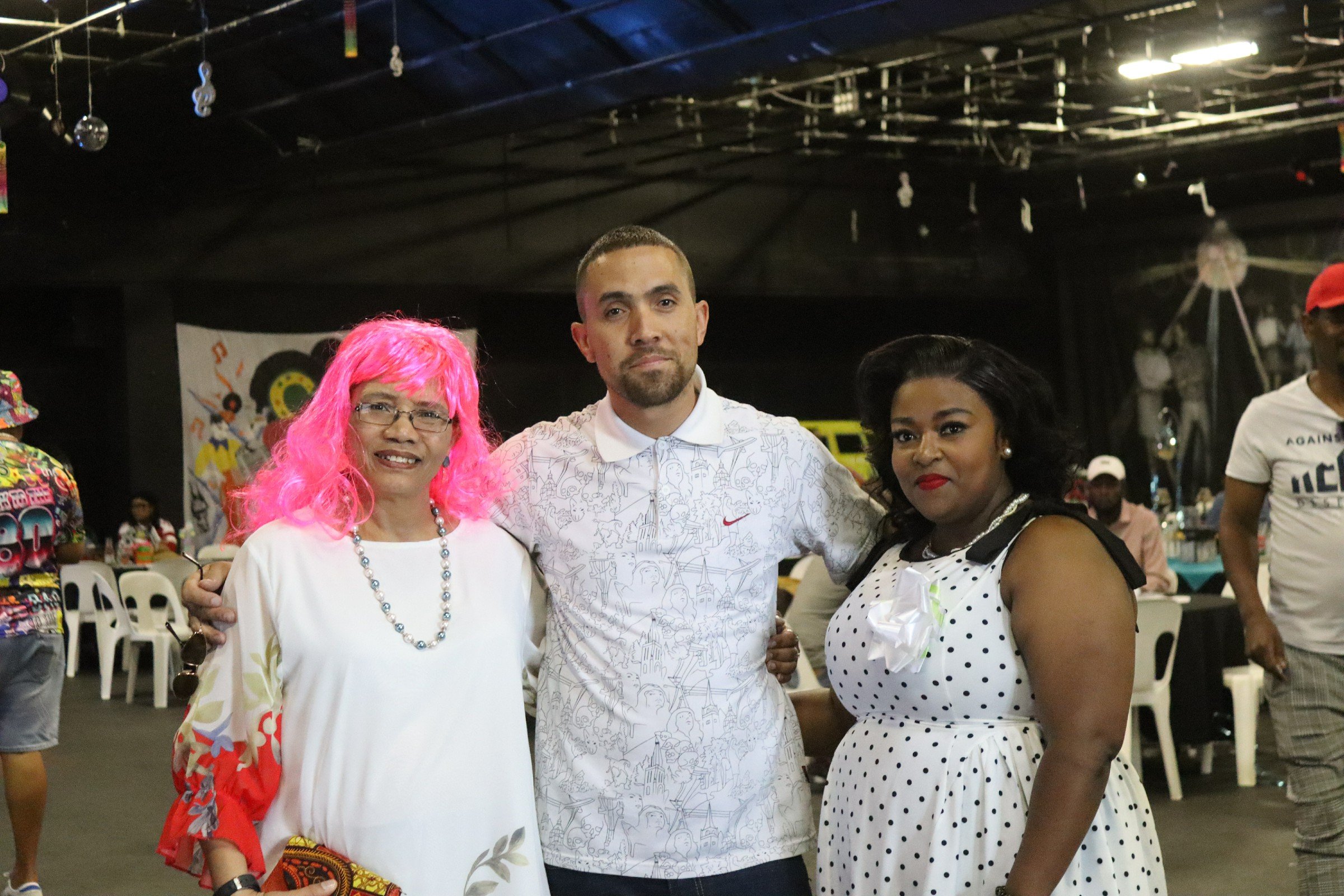 Three people standing together at an indoor event, smiling at the camera. The woman on the left has pink hair, glasses, and is wearing a white dress with colorful patterns and jewelry. The man in the middle has short hair and a beard, wearing a white