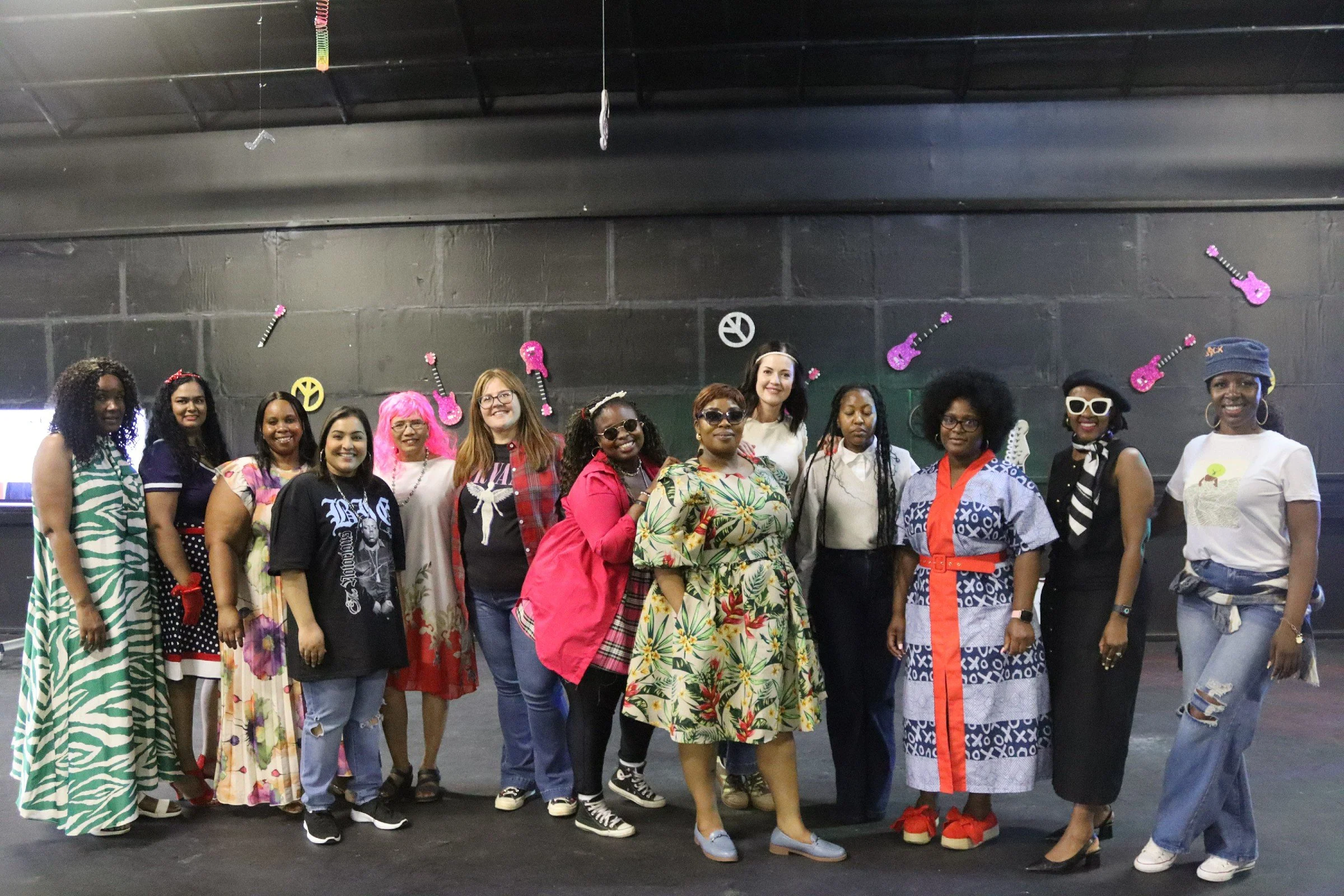 A group of diverse women standing together indoors against a black wall decorated with pink and yellow guitar and peace sign cutouts. They are smiling and dressed in colorful, patterned clothing.