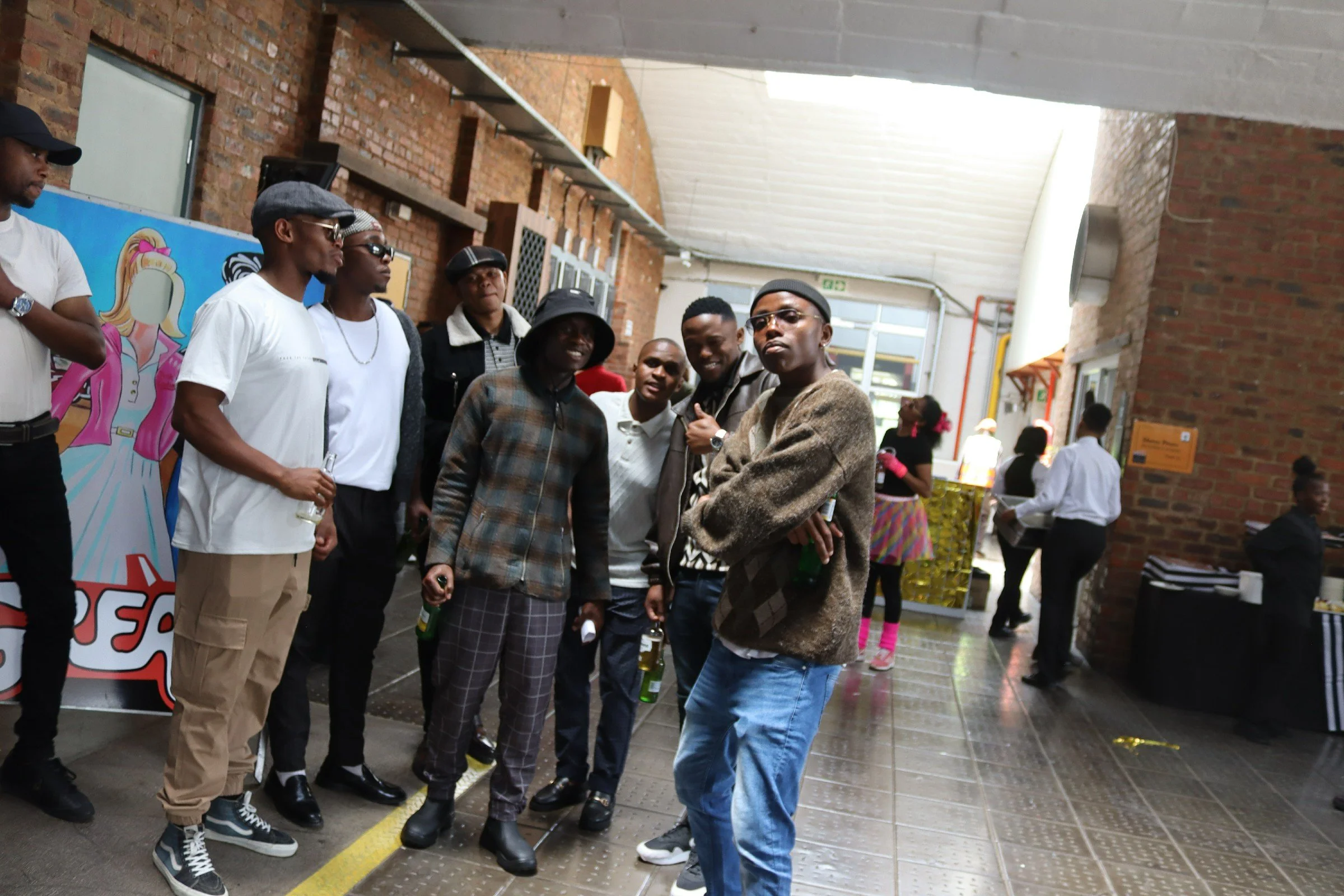 A group of people gathered indoors near brick walls, some wearing sunglasses and hats, with a colorful cartoon poster and other individuals in the background.