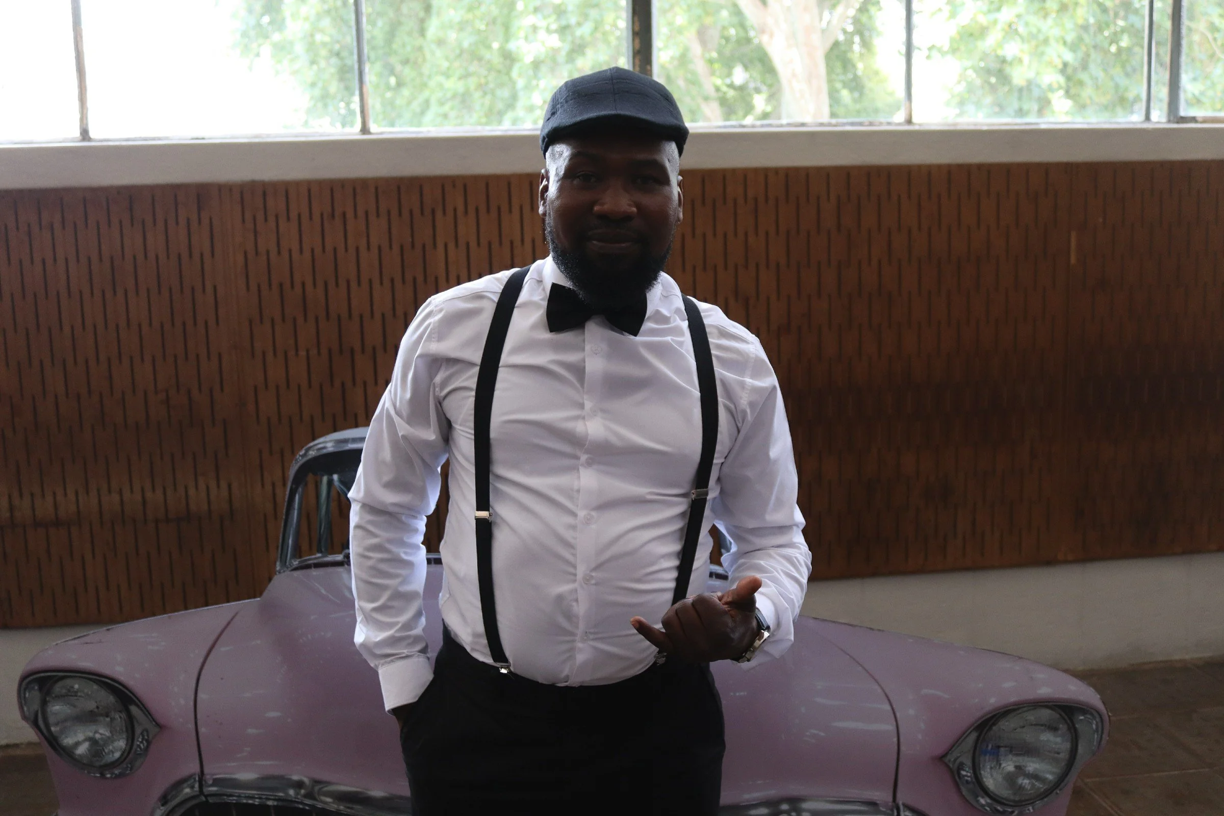A man dressed in a white shirt, black suspenders, and a black bow tie, wearing a black cap, standing in front of a pink vintage car inside a building with wooden panel walls and large windows showing green trees outside.