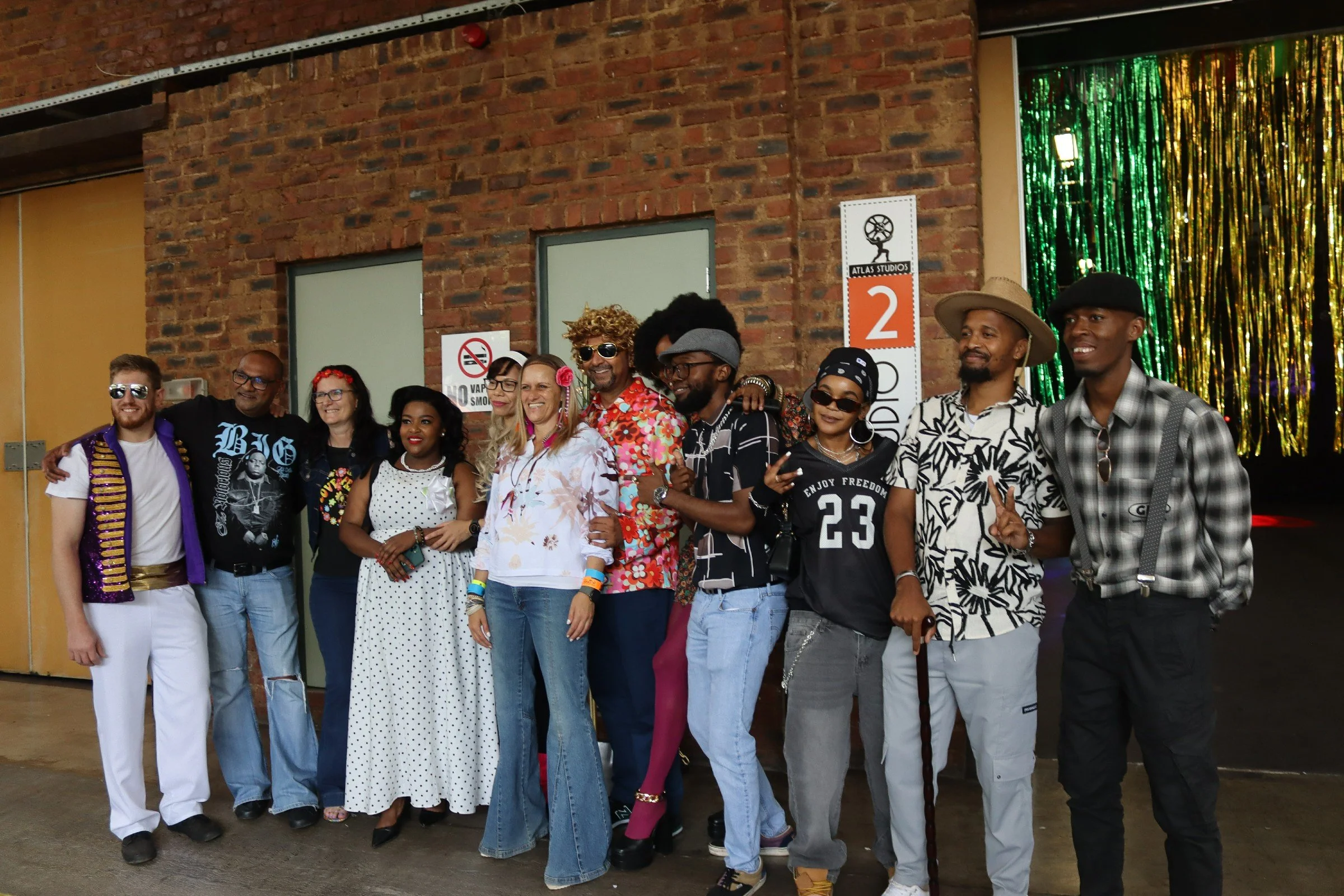 A diverse group of people in colorful clothing posing together for a photo in front of a brick wall and a shiny curtain backdrop at an indoor event.