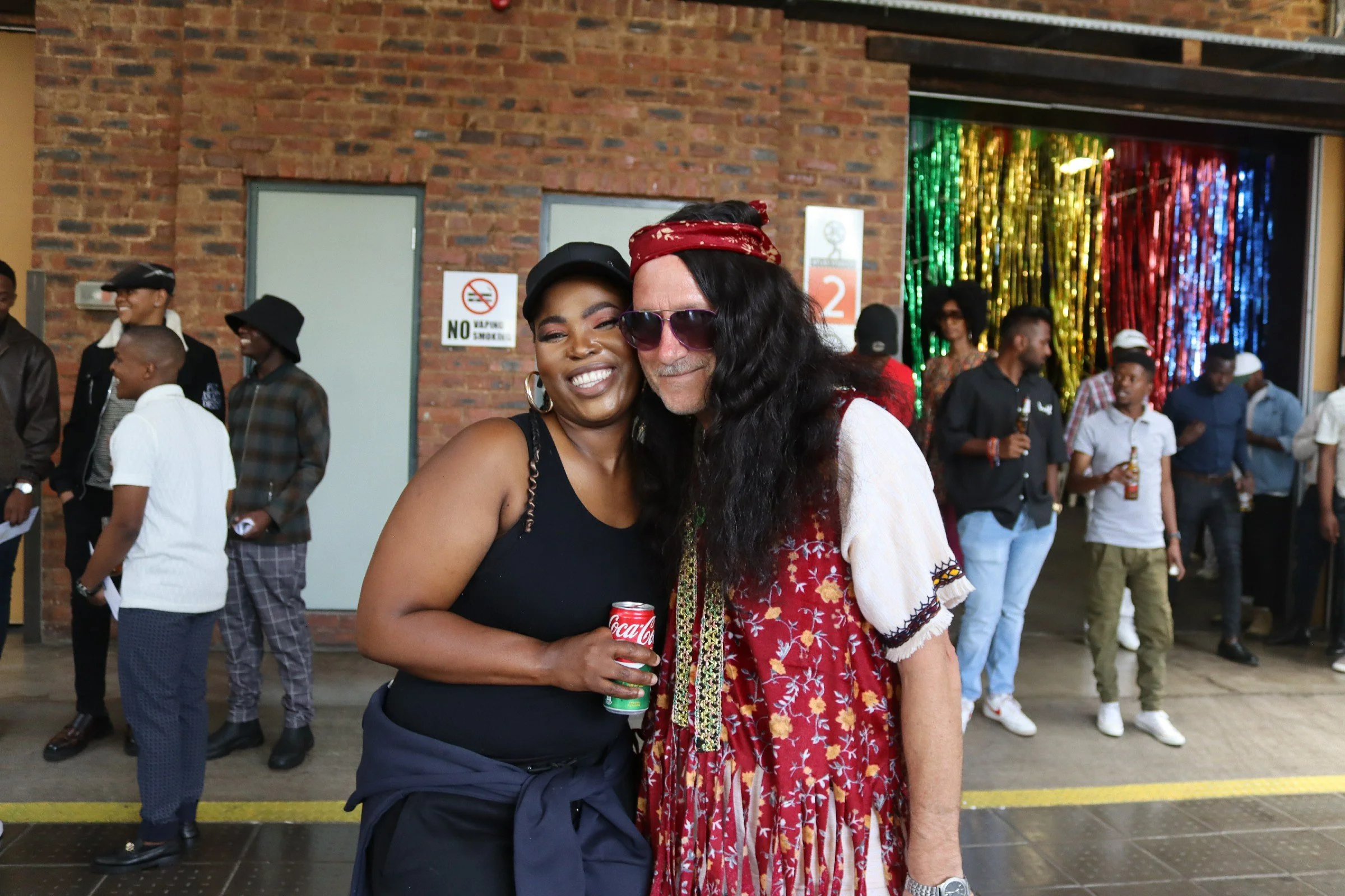 Two women smiling and hugging at a social gathering. One woman is wearing a black tank top and holding a Diet Coke, the other is dressed in a colorful, patterned dress with a red headscarf and sunglasses. They are standing in front of a brick wall wi