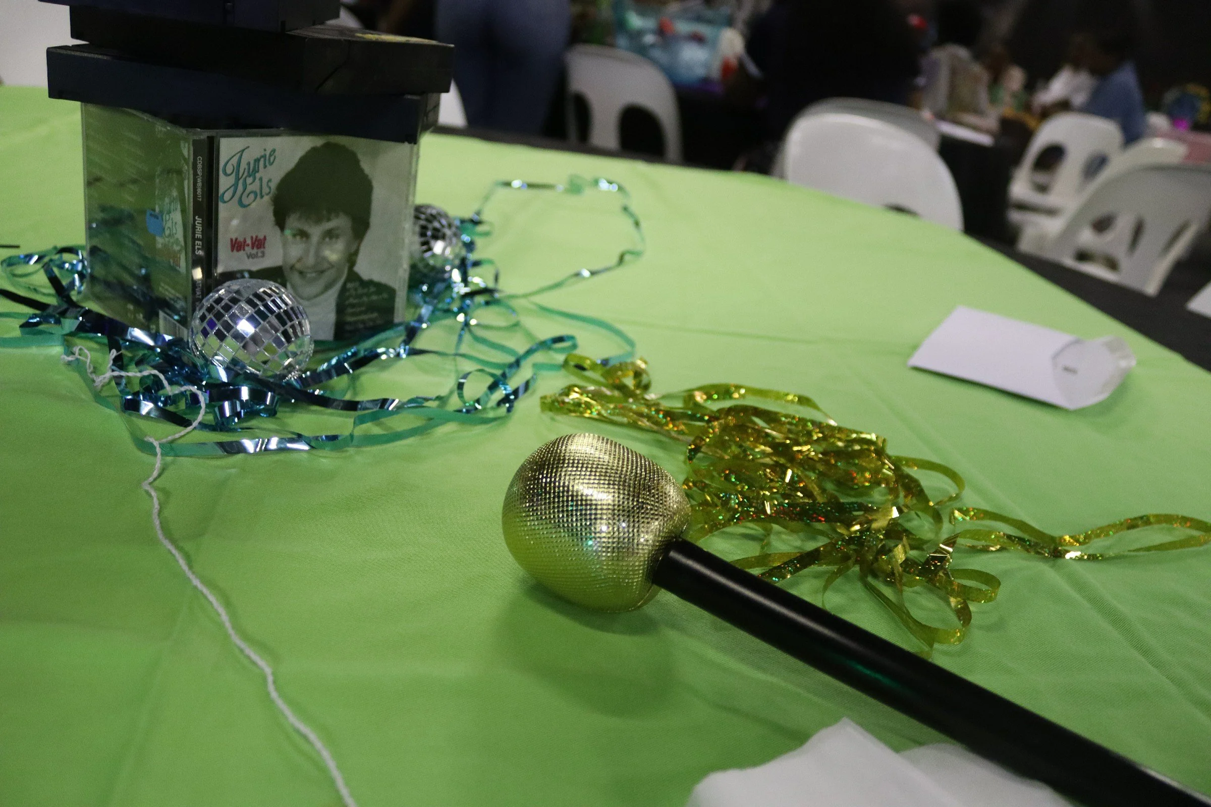 Table decorated with silver and gold tinsel, small disco ball, gold ornament, black microphone, and a flyer with a woman's face and text, on a green tablecloth in an event hall.