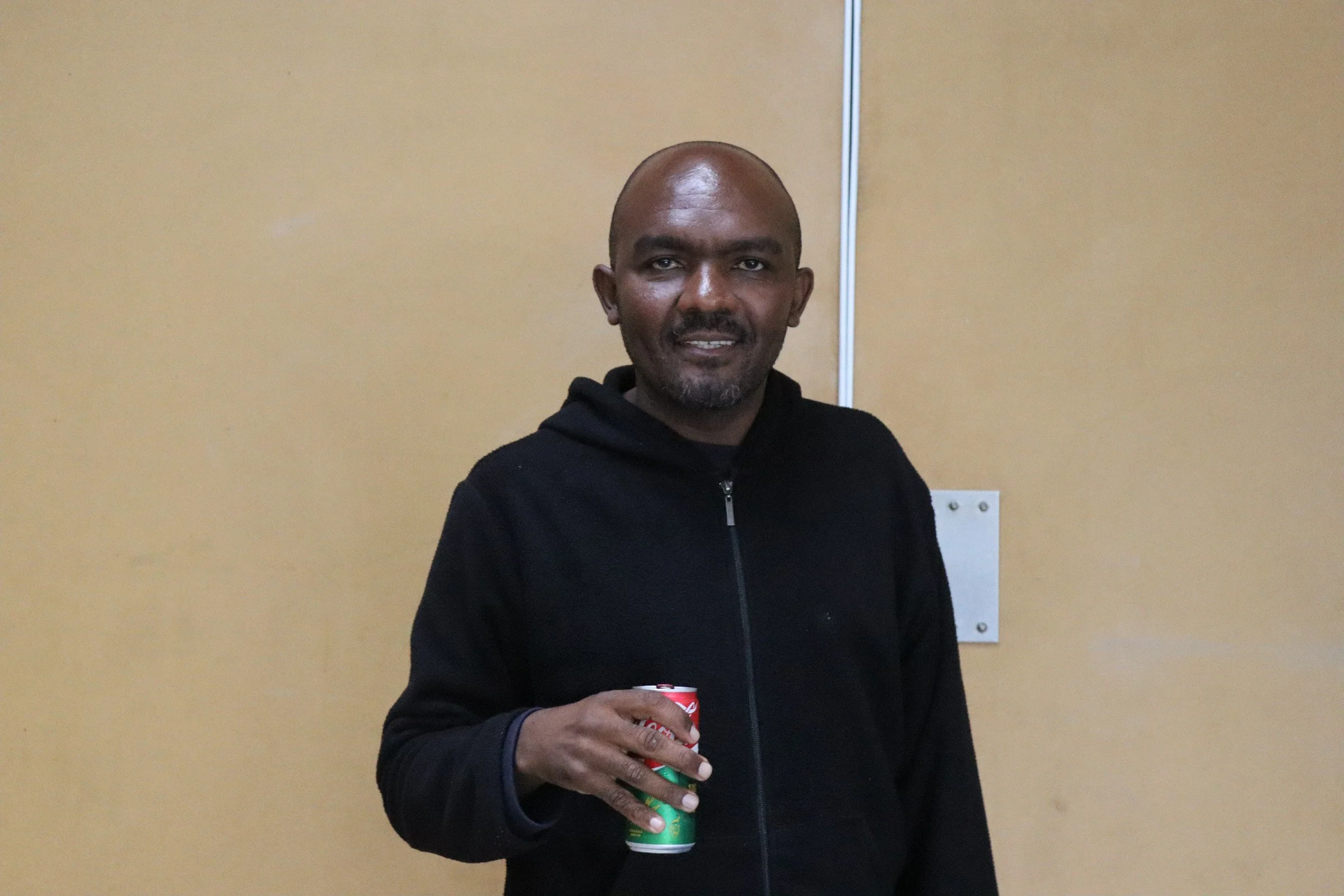 A man with a shaved head and goatee, wearing a black zip-up hoodie, is standing in front of a beige wall, holding a can of soda in his right hand and smiling slightly.