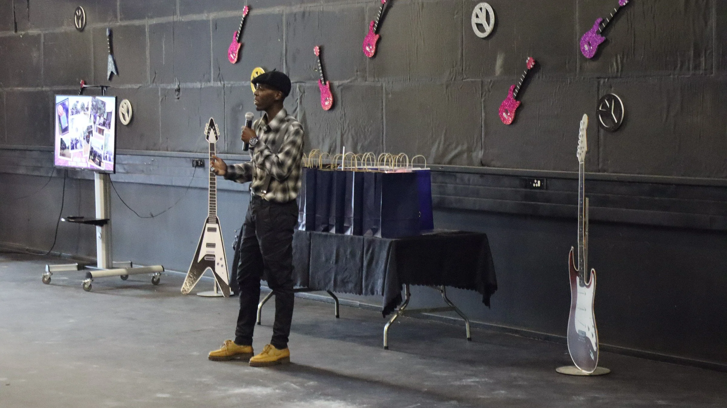 A young man standing on a stage giving a presentation, holding a microphone. Behind him, there is a black wall decorated with pink and white glittery guitar cutouts and black peace sign symbols. To his left, a flat-screen monitor displays photos, and