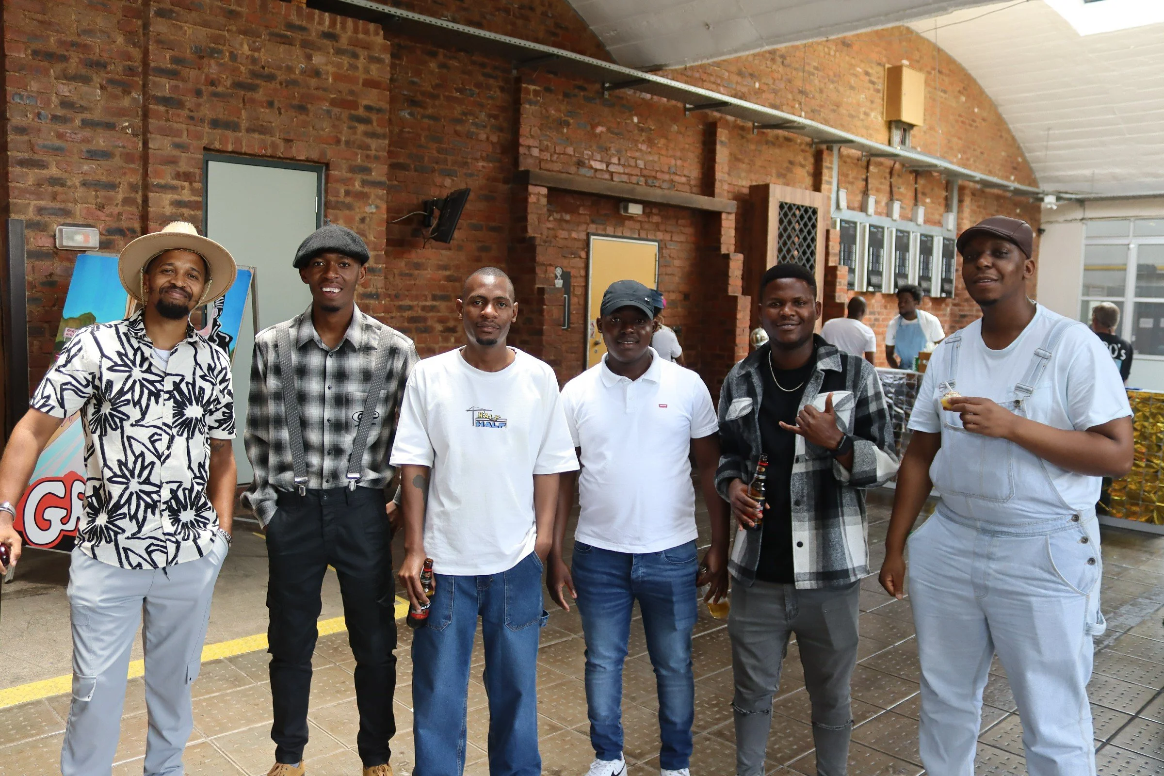 A group of seven people standing inside a brick-walled building, posing for a photo. They are wearing casual clothing and some are holding drinks, with a mixture of facial expressions and gestures. The background shows a few other people and some ven