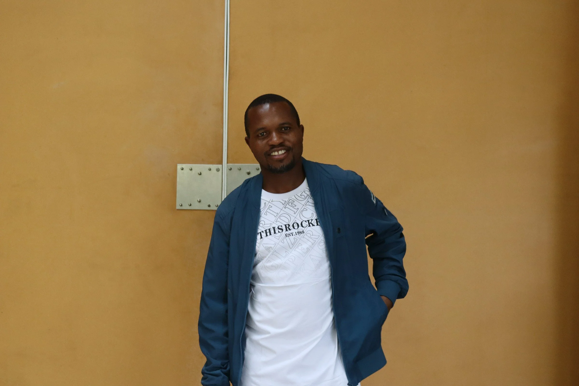 A man standing in front of a beige wall, smiling, wearing a white T-shirt and a blue jacket.