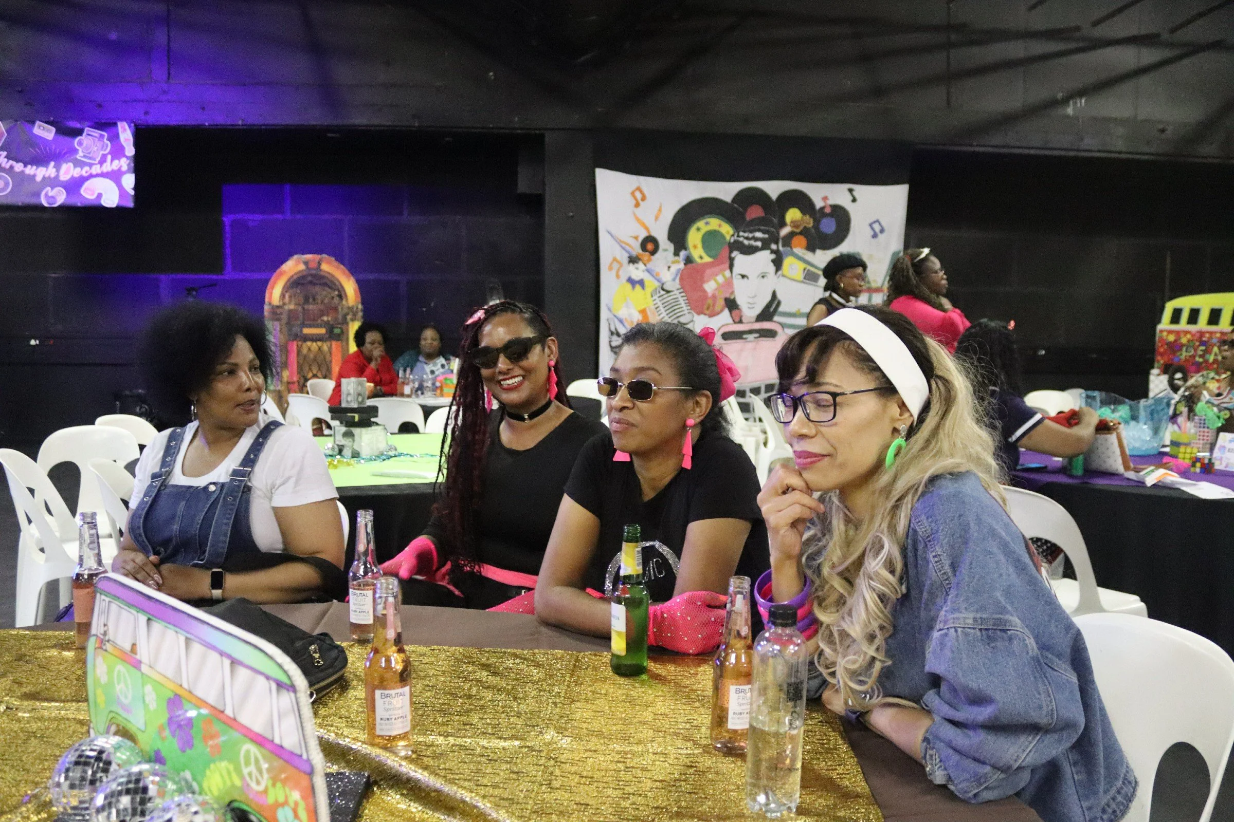 A group of women sitting at a table during a celebration or event with colorful decorations, drinks, and gifts.