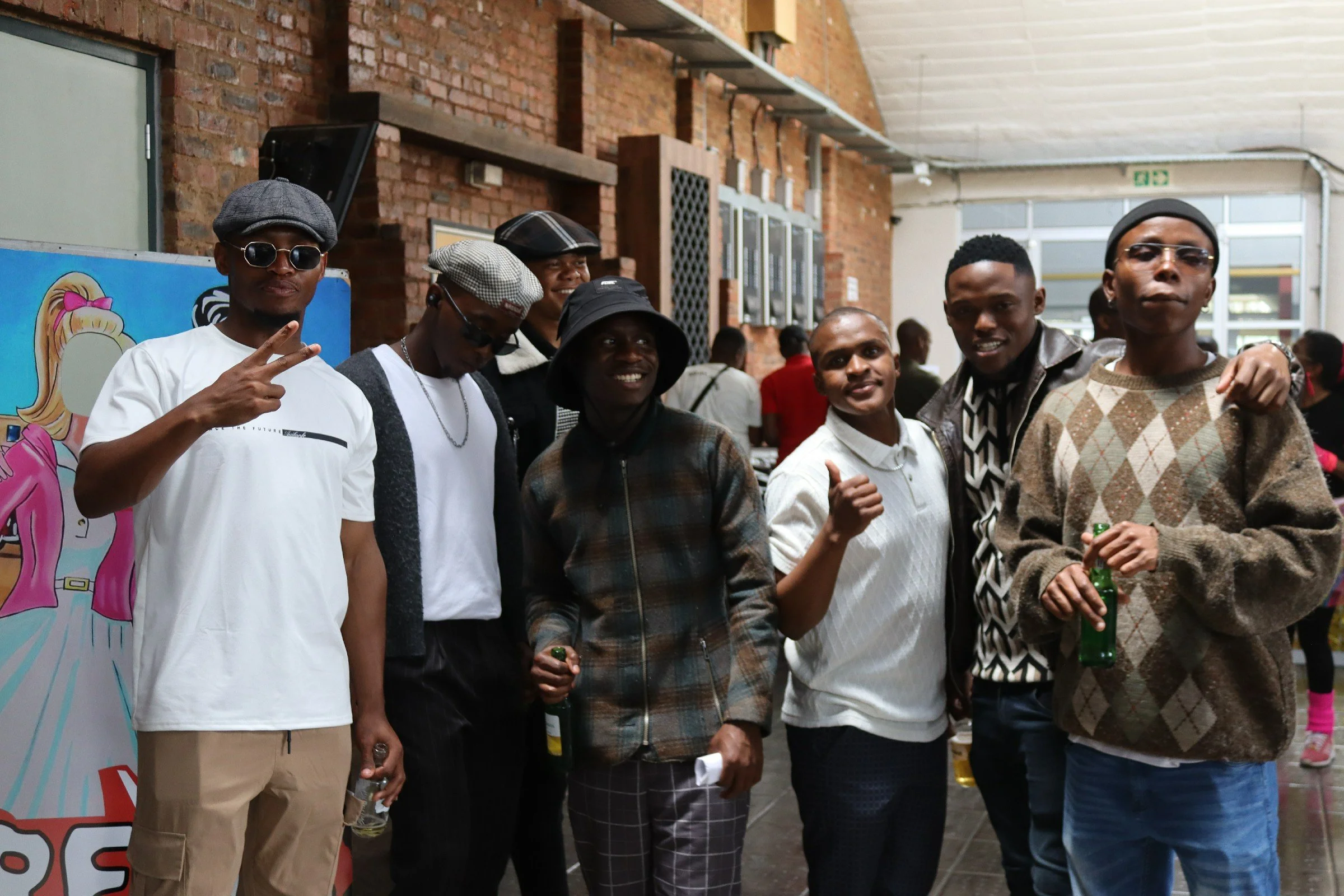 A group of seven young men standing closely together inside a building with brick walls. Some are wearing sunglasses and hats, and they are smiling or posing for the photo. The background includes a colorful poster and industrial-looking elements lik