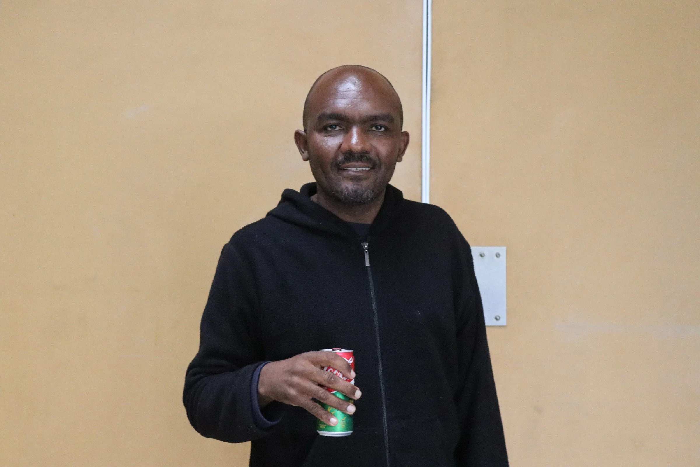 A man with a shaved head and goatee wearing a black hoodie holding a can of soda.