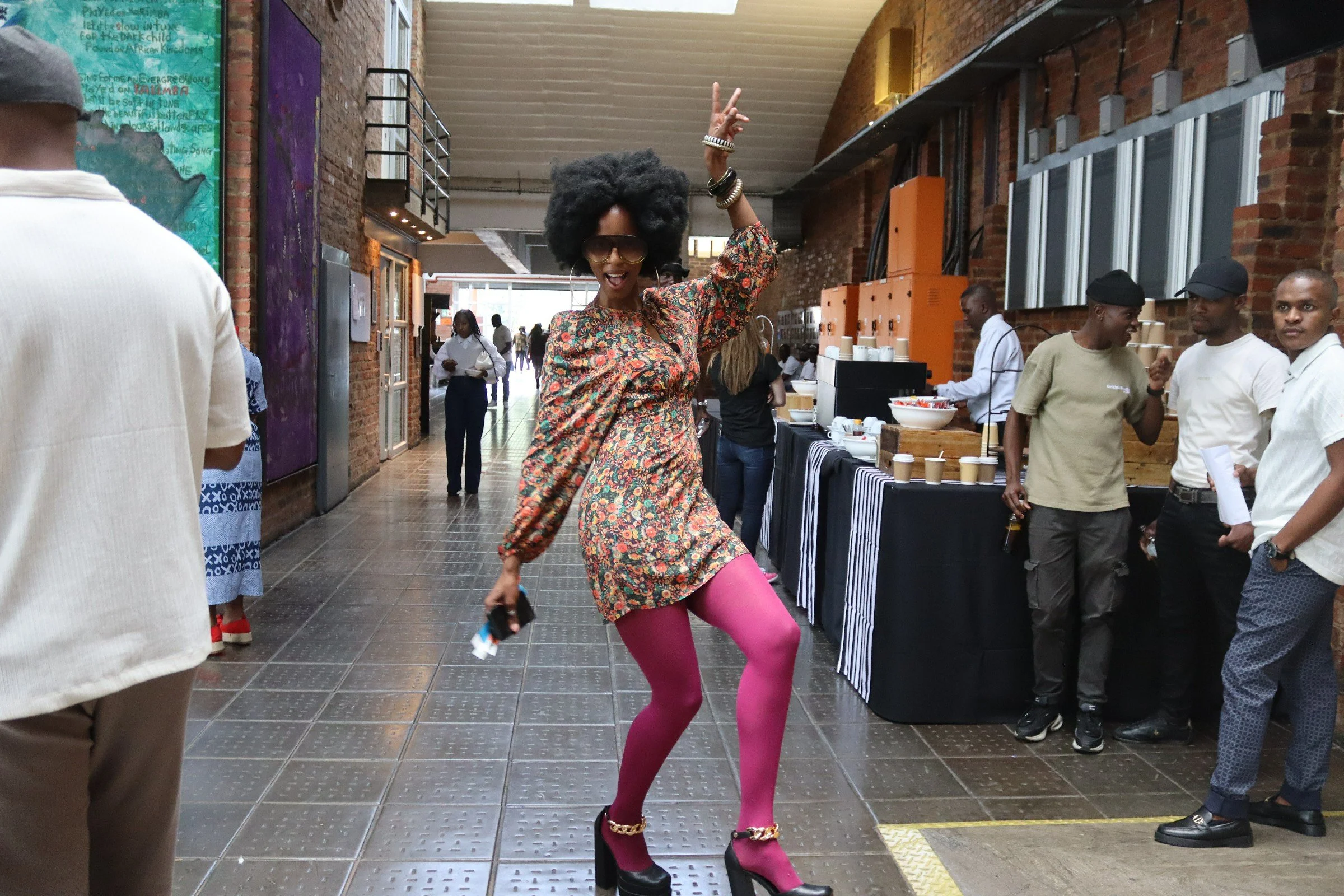 A woman with a large afro hairstyle, wearing sunglasses, a colorful patterned dress, bright pink tights, and high-heeled shoes, dancing happily in a busy indoor setting with brick walls and people in the background.