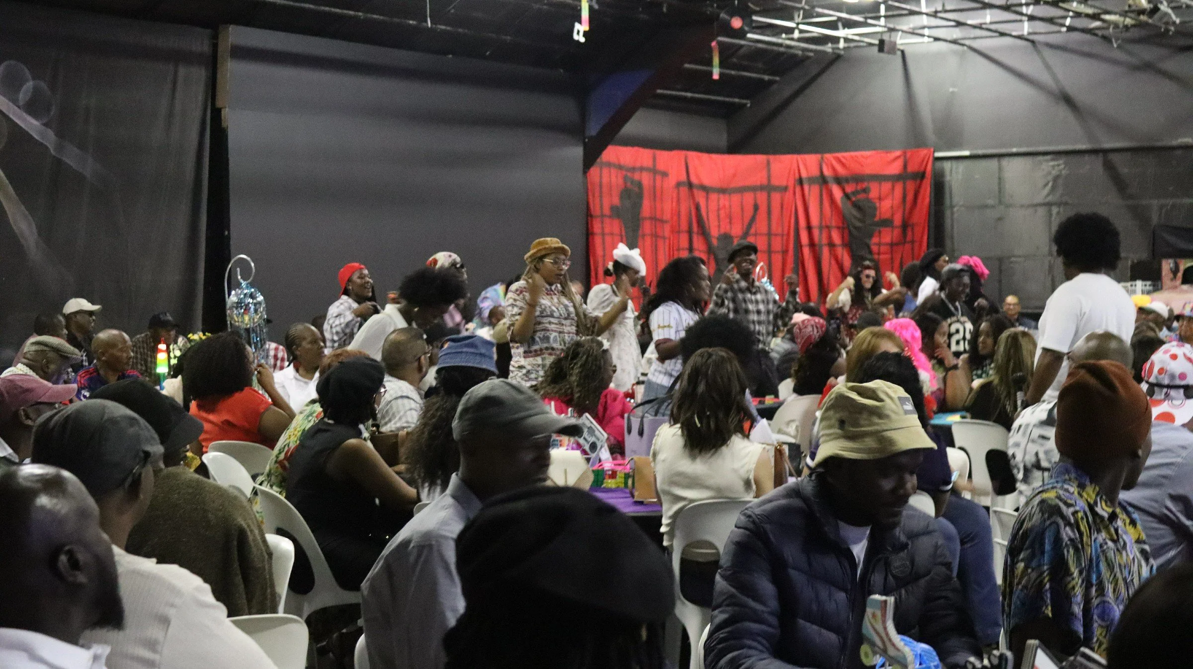 A large gathering of people seated indoors, many wearing colorful clothing and hats, with some standing and appearing to be participating in a celebration or event, with a red curtain or backdrop and dark walls in the background.