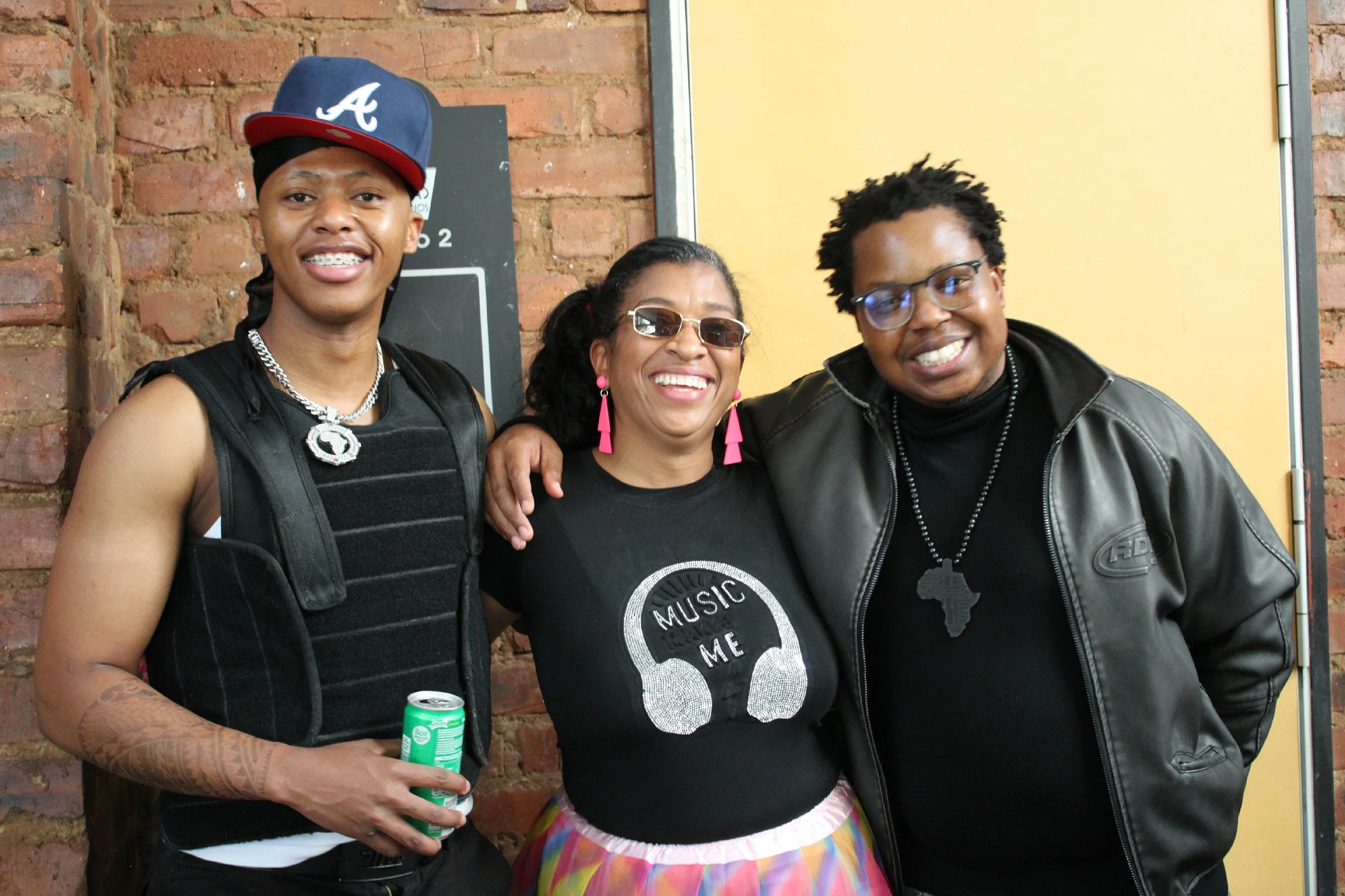 Three smiling people, two men and a woman, standing together in front of a brick wall and yellow door. The man on the left is wearing a backwards baseball cap, a black body armor vest, and holding a canned drink. The woman in the middle has glasses, 