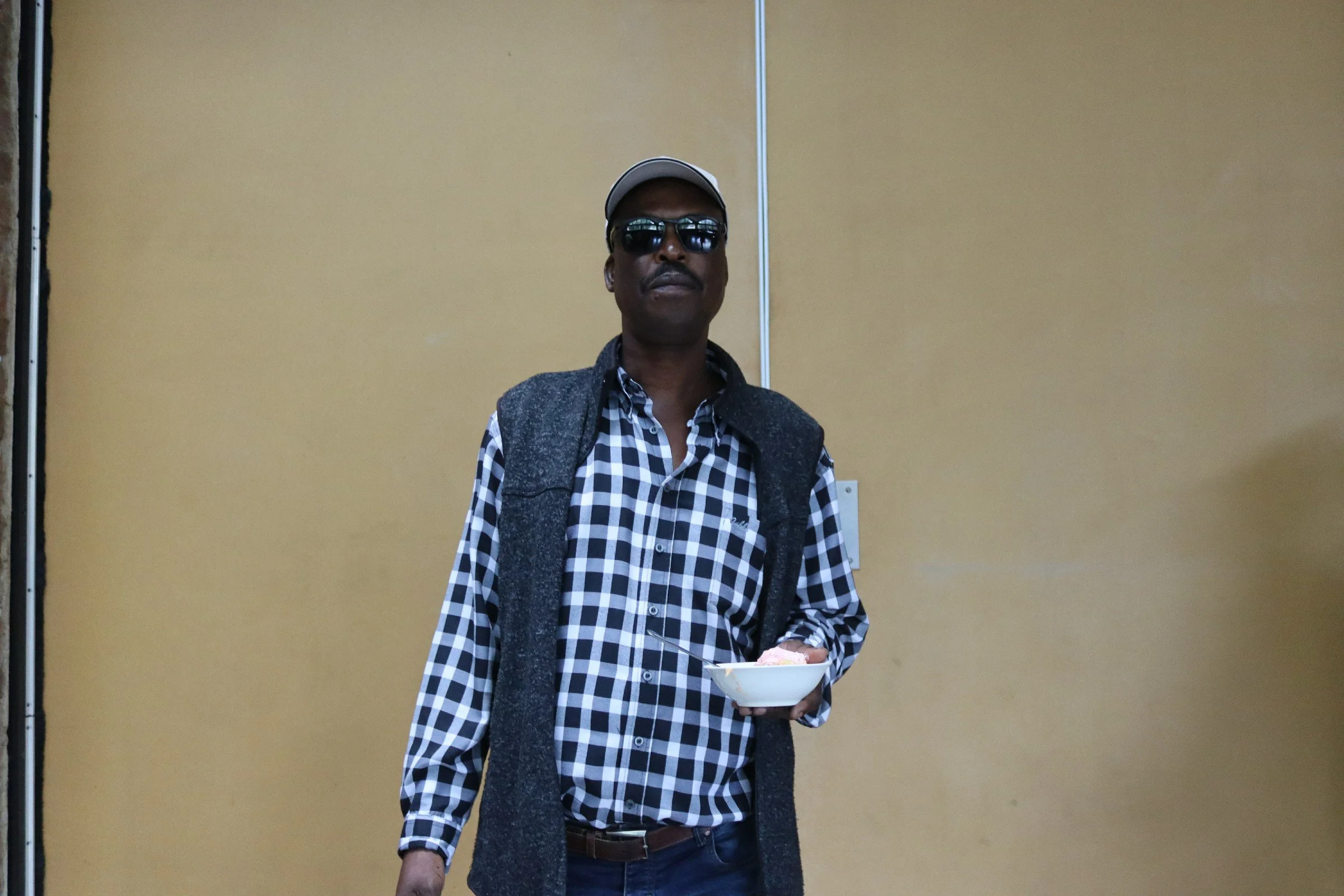 A man wearing black sunglasses, a gray cap, a checkered shirt, and a dark gray vest, holding a white bowl, stands in front of a plain yellow wall.