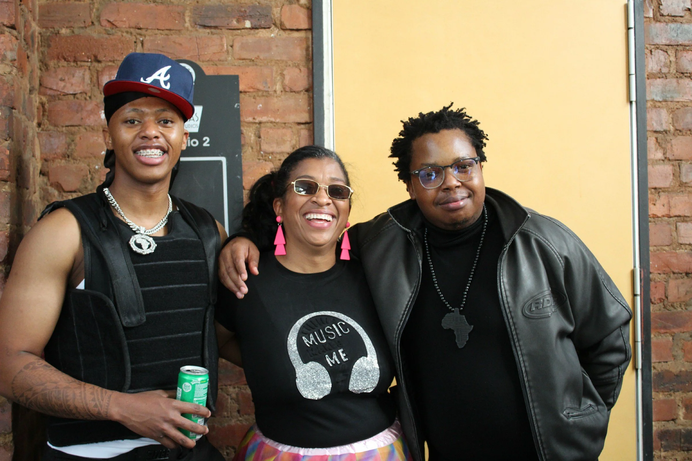Three people standing together indoors, smiling at the camera, with brick walls and a yellow partition in the background. The person on the left wears a blue and red cap, a tattooed arm, and a chest protector. The person in the middle wears glasses, 