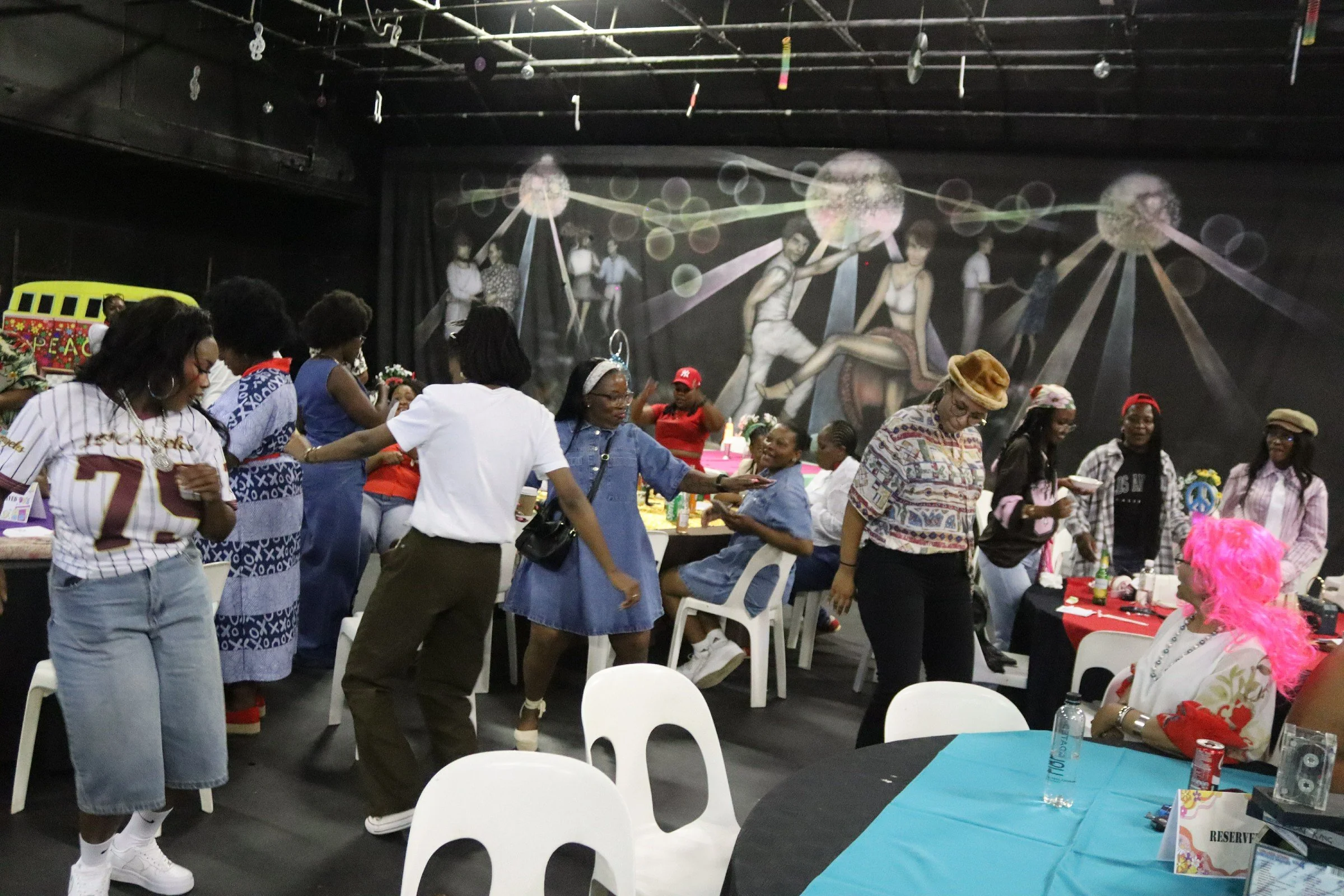 A group of people gathered at a party or event, some standing and others sitting at tables with blue tablecloths, in a room with a black ceiling and a dark wall mural featuring abstract figures. The room has chairs, drinks, and decorations, with some