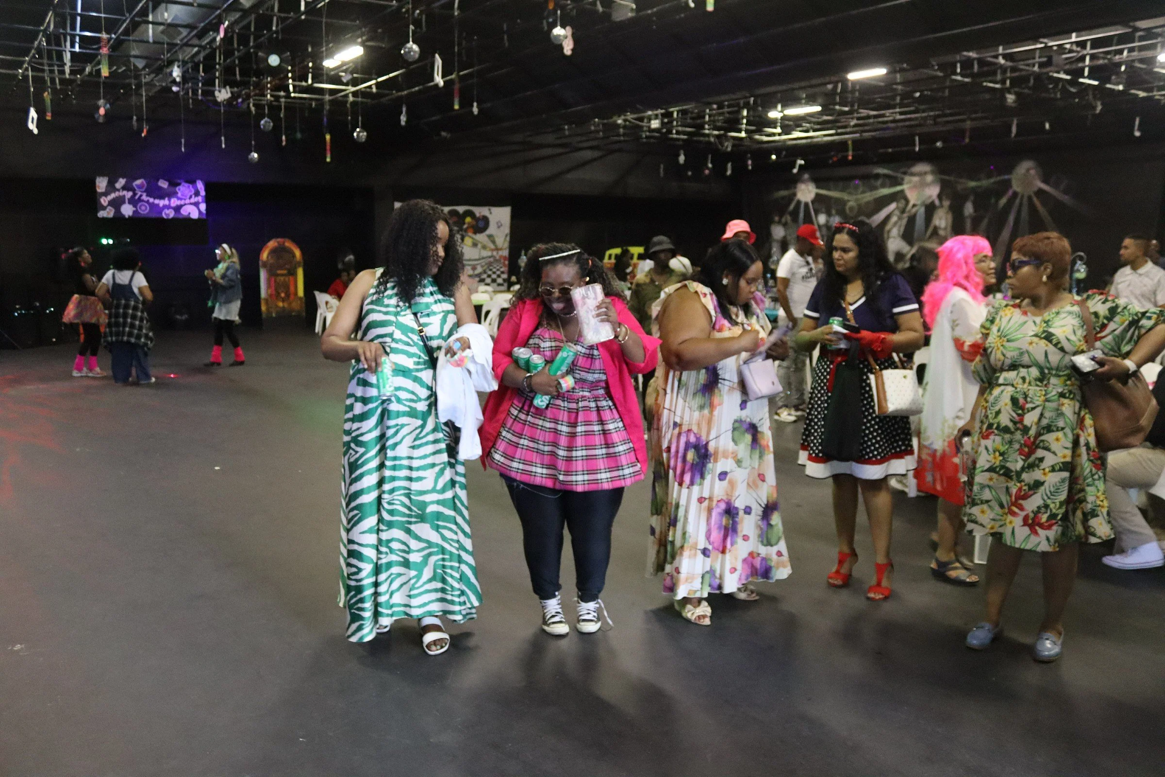 A group of women dressed in colorful dresses are standing and walking in an indoor venue with dark lighting and hanging decorations, possibly at a social event or gathering.