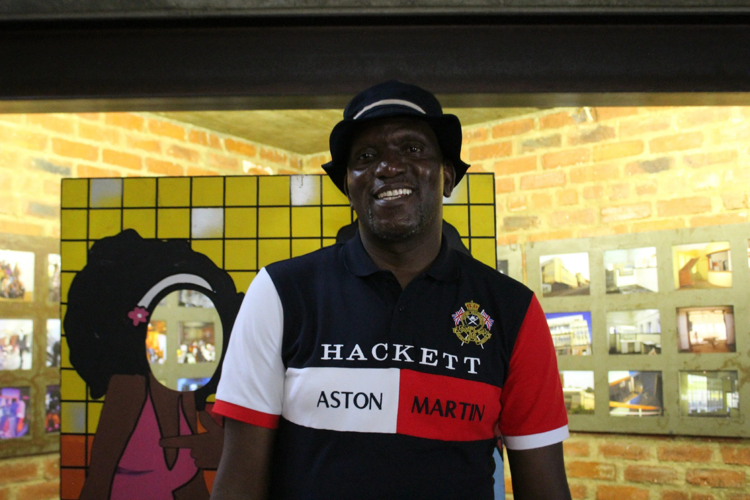 A man smiling, wearing a black hat and a patterned polo shirt with the words "HACKETT ASTON MARTIN" on it, stands in front of a yellow and black backdrop with a pink cartoon horse figure and brick wall behind him.